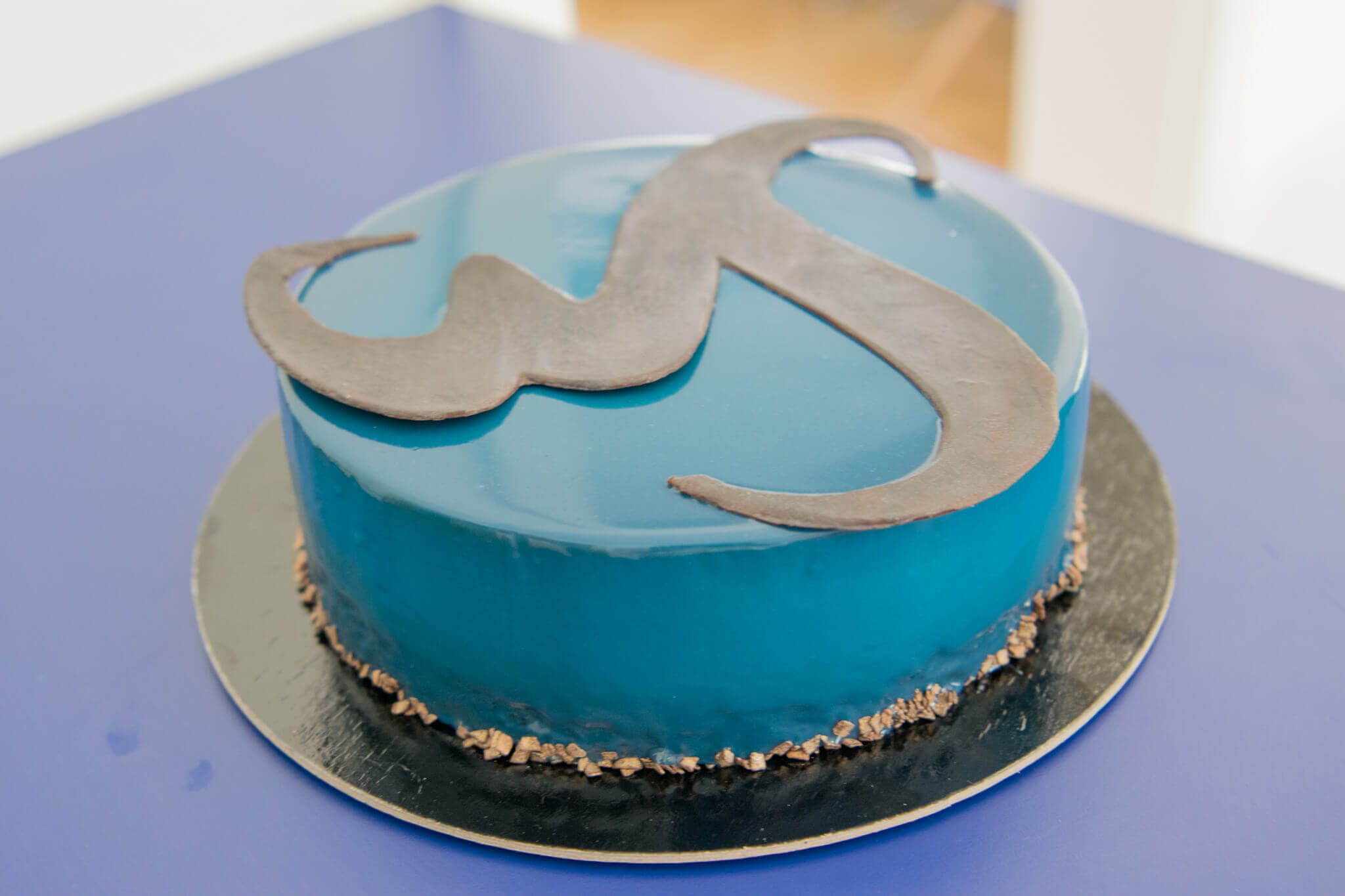 Blaue Torte mit glänzendem Überzug und schwarzer Schokoladendekoration auf einem Tisch.