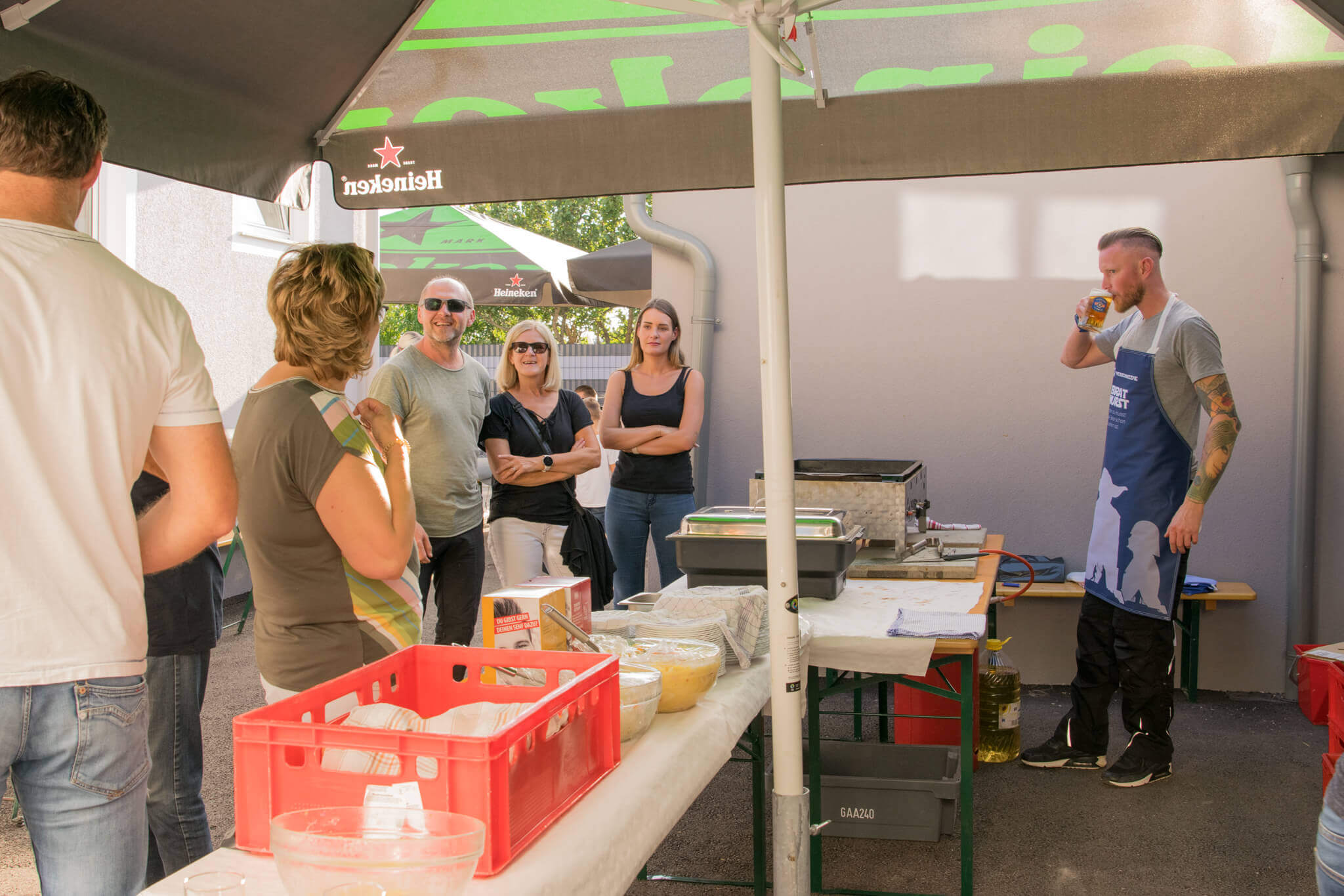Grillfest im Freien: Menschen unter Heineken-Schirmen, ein Mann trinkt Bier neben einem Grill.