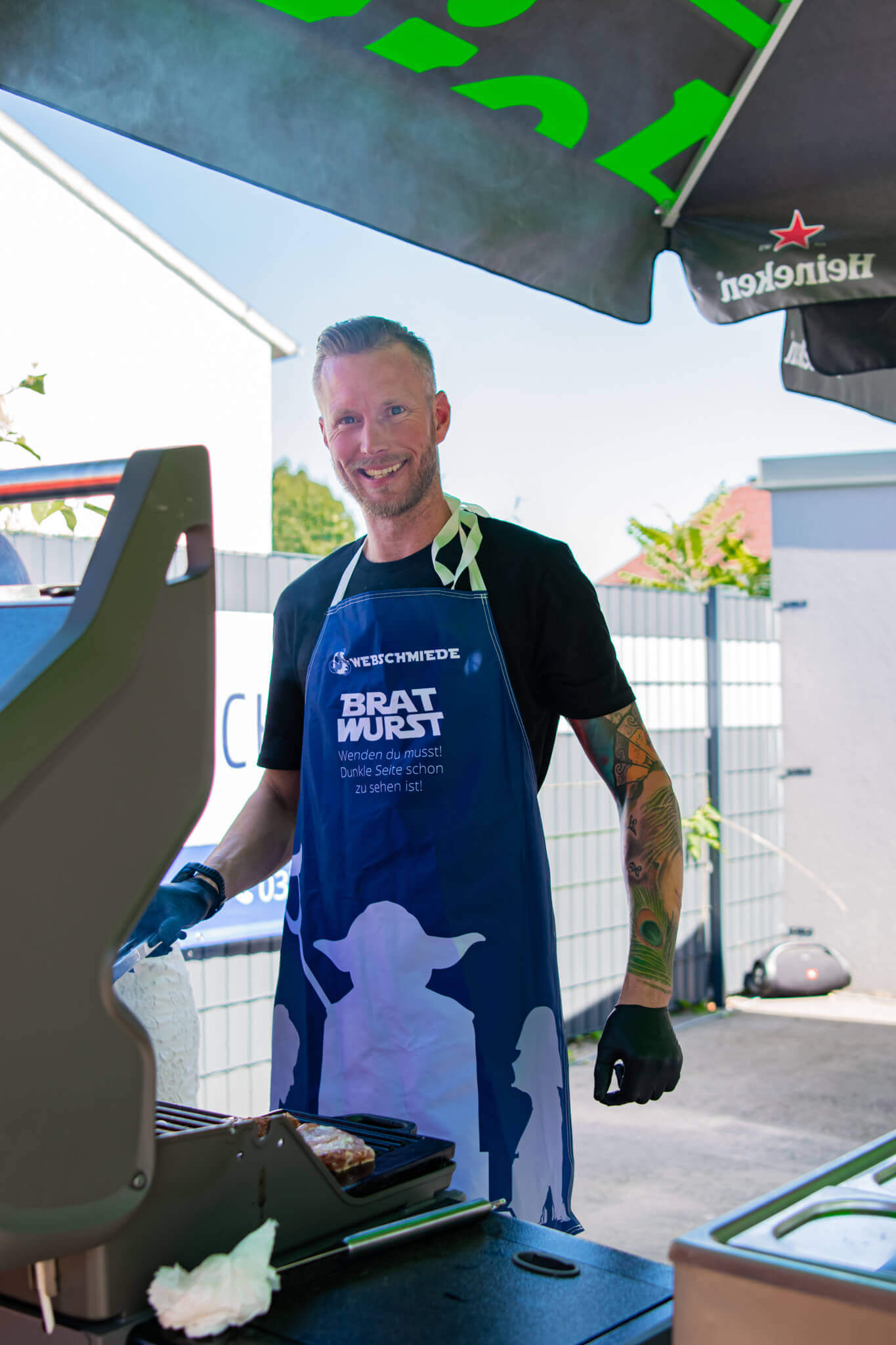 Grillmeister in Schürze mit witzigem Spruch steht fröhlich am Grill im Garten unter einem Sonnenschirm.