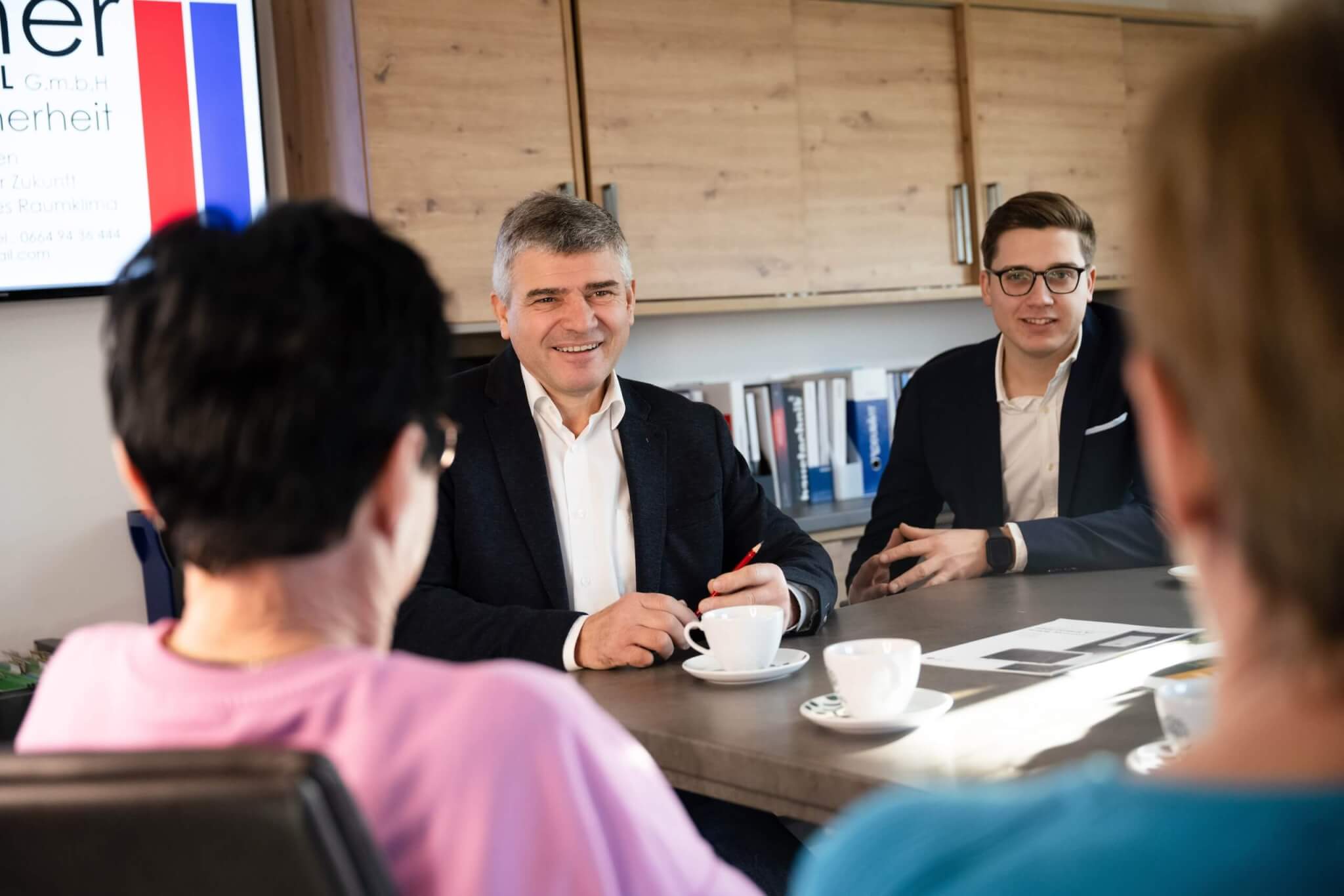 Besprechung im Büro: Vier Personen diskutieren lächelnd am Konferenztisch mit Kaffeetassen und Unterlagen in moderner Umgebung.