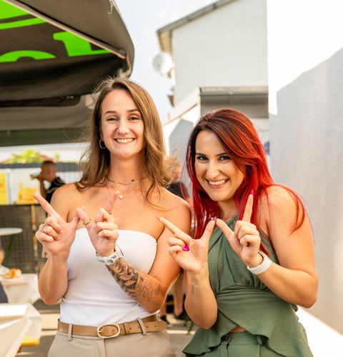 Zwei lächelnde Frauen bei einem Event im Freien, die in die Kamera zeigen.