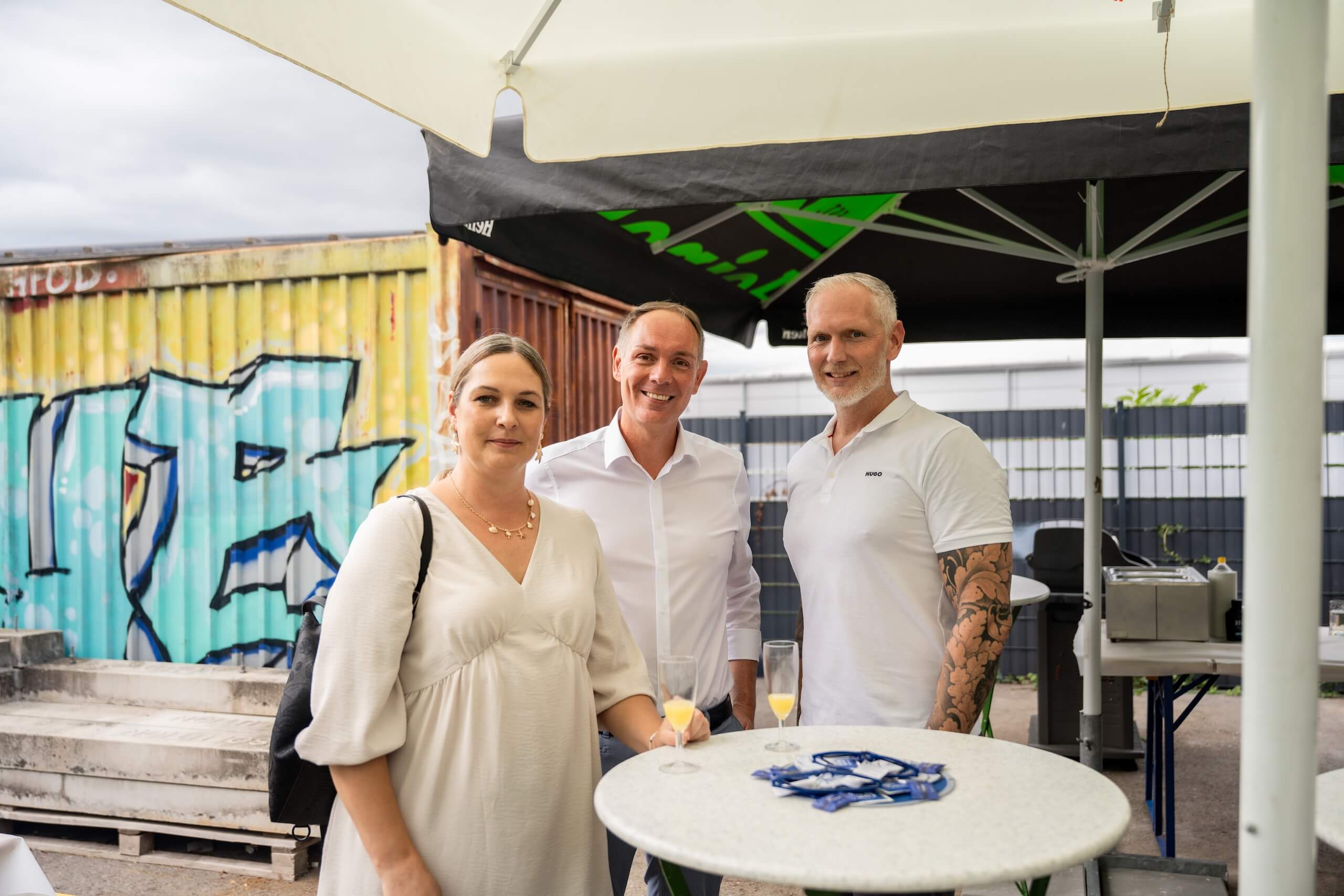Personen beim Networking-Event unter einem Sonnenschirm mit Getränken, Graffiti im Hintergrund.