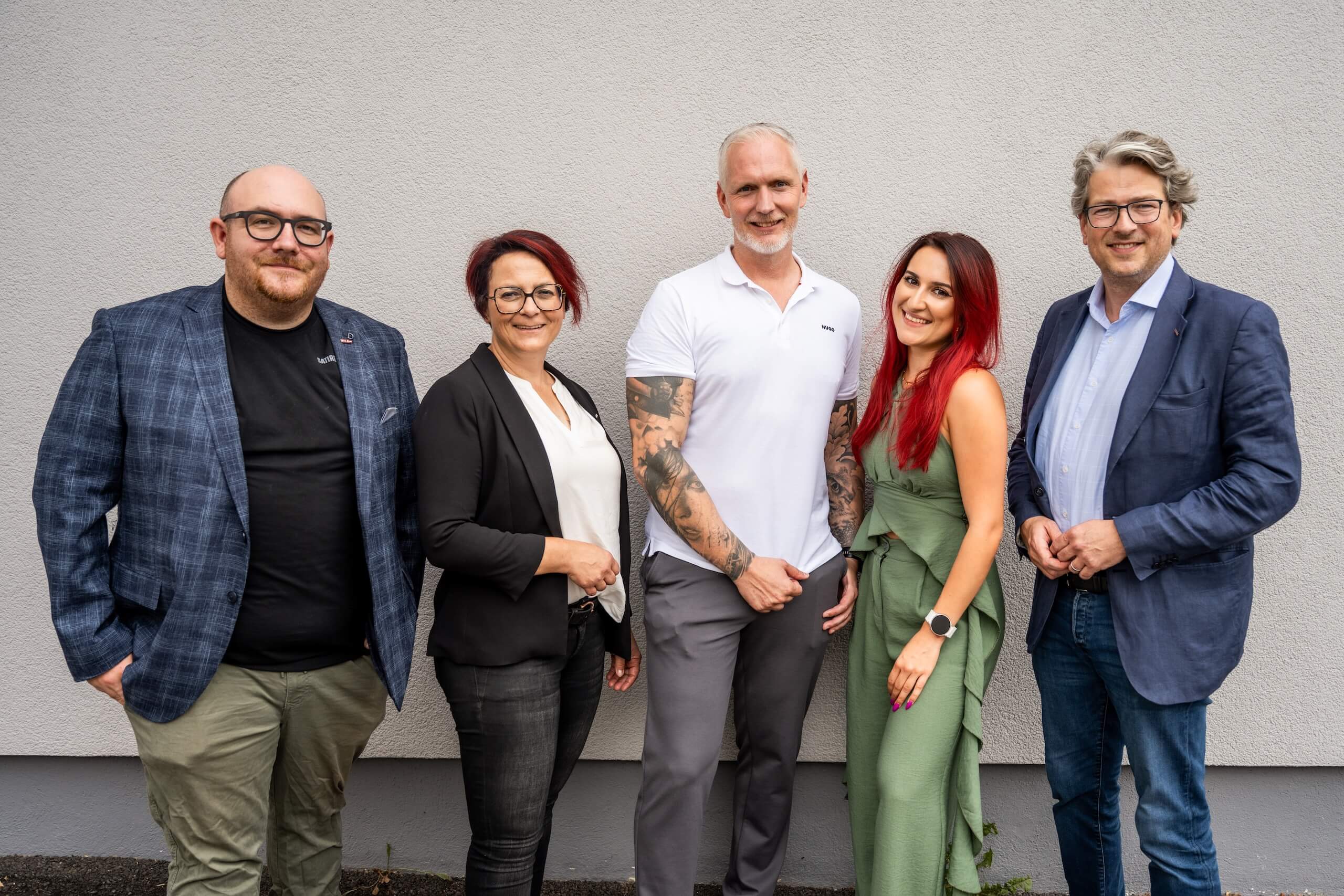 Gruppe von fünf lächelnden Personen posiert vor einer grauen Wand. Business-Kleidung, diverse Aufmachung. Teamkonzept.