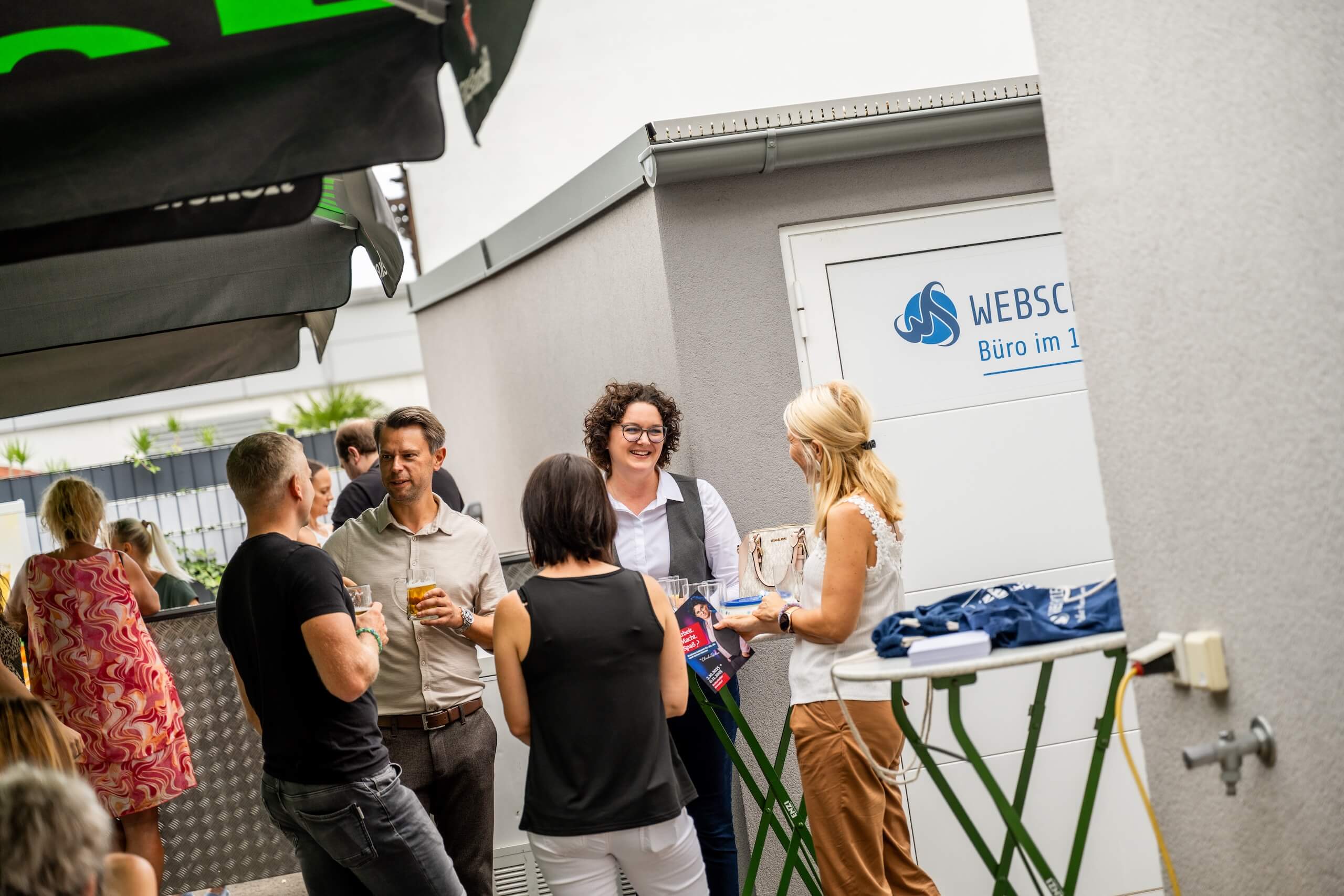 Personen unterhalten sich bei einem Networking-Event im Freien vor einem Büro mit Webschmiede-Logo.