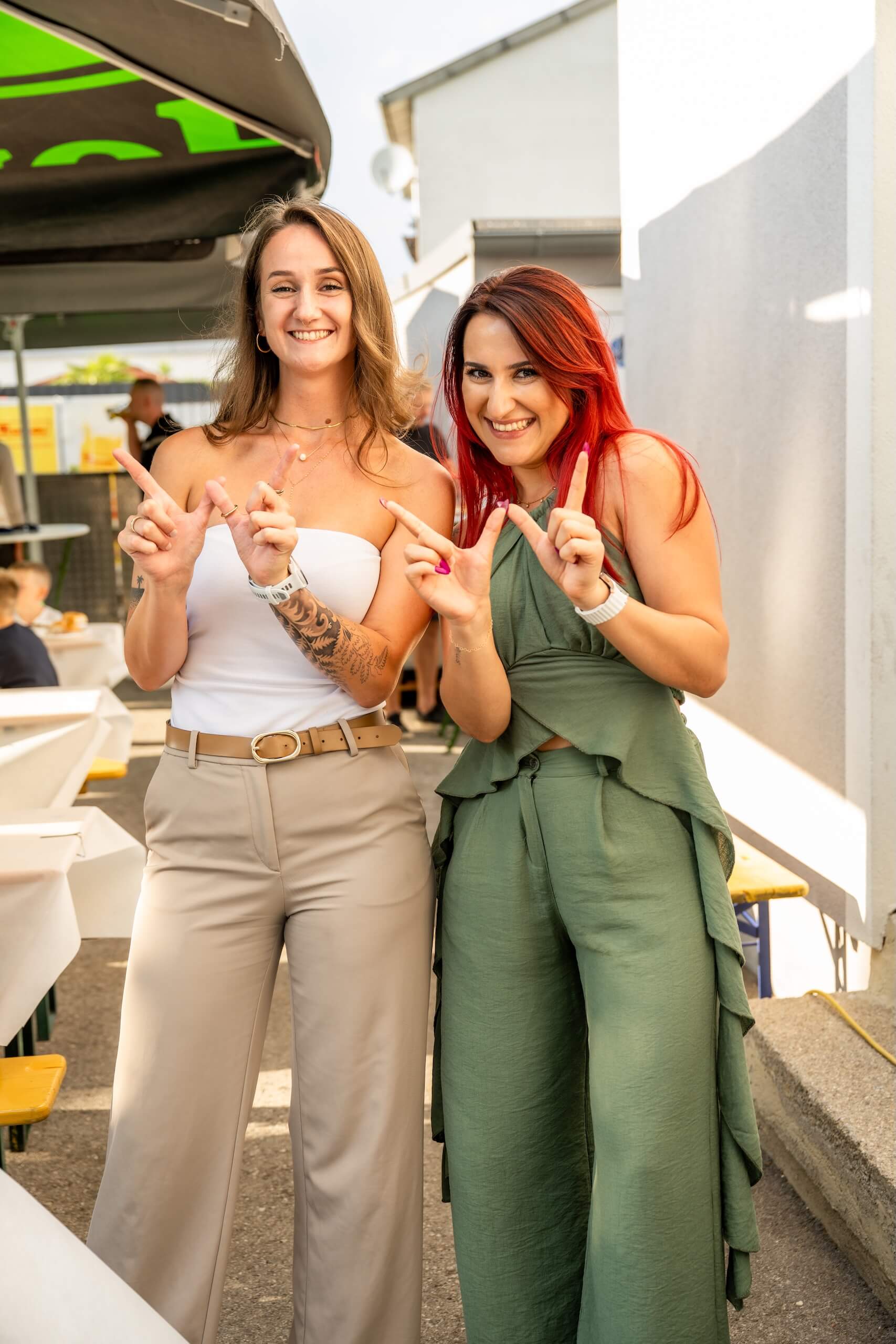 Zwei lächelnde Frauen im Freien posieren fröhlich für ein Foto und zeigen Handgesten.