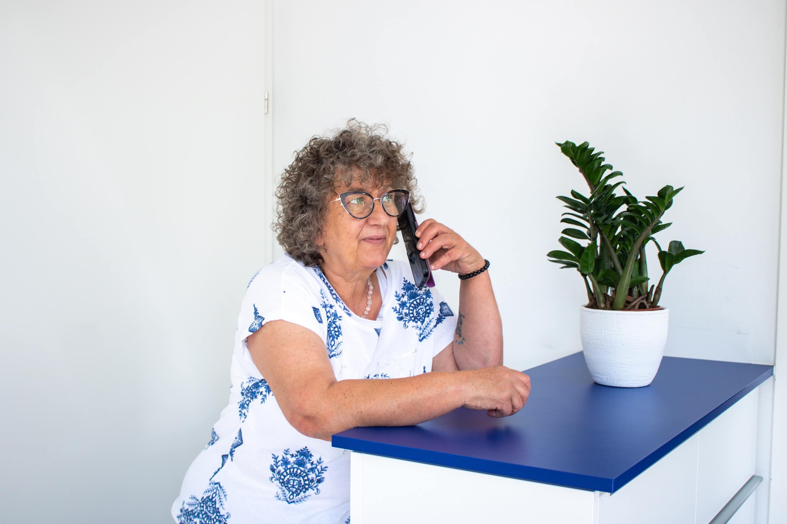 Ältere Frau telefoniert lächelnd am Tisch mit Topfpflanze, trägt Brille und blau-weißes Musterkleid.