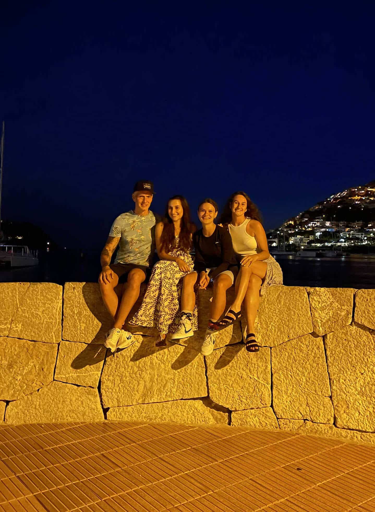 Vier Personen sitzen abends auf einer Mauer am Wasser, mit beleuchtetem Hügel im Hintergrund.
