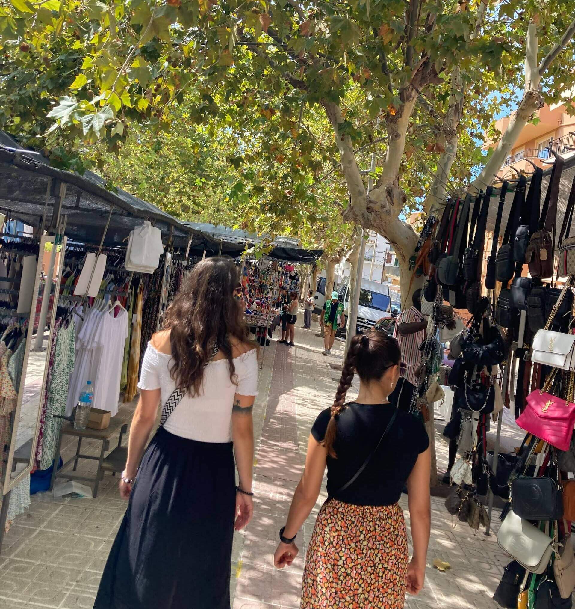 Zwei Personen schlendern über einen belebten, schattigen Straßenmarkt mit Ständen voller Kleidung und Taschen.