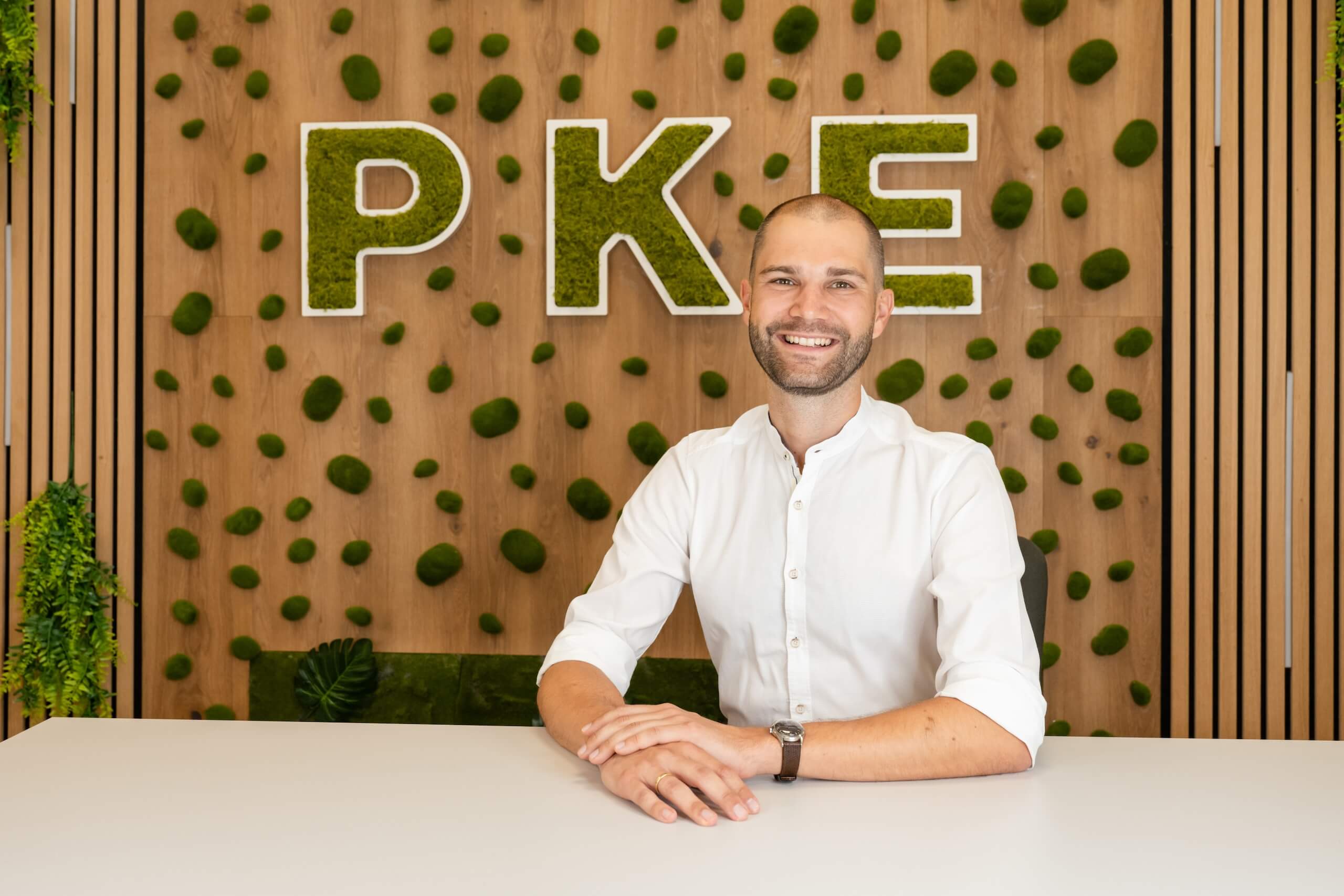 Mann sitzt lächelnd vor einer mit Moos dekorierten Wand mit PKE-Logo in einem modernen Büro.