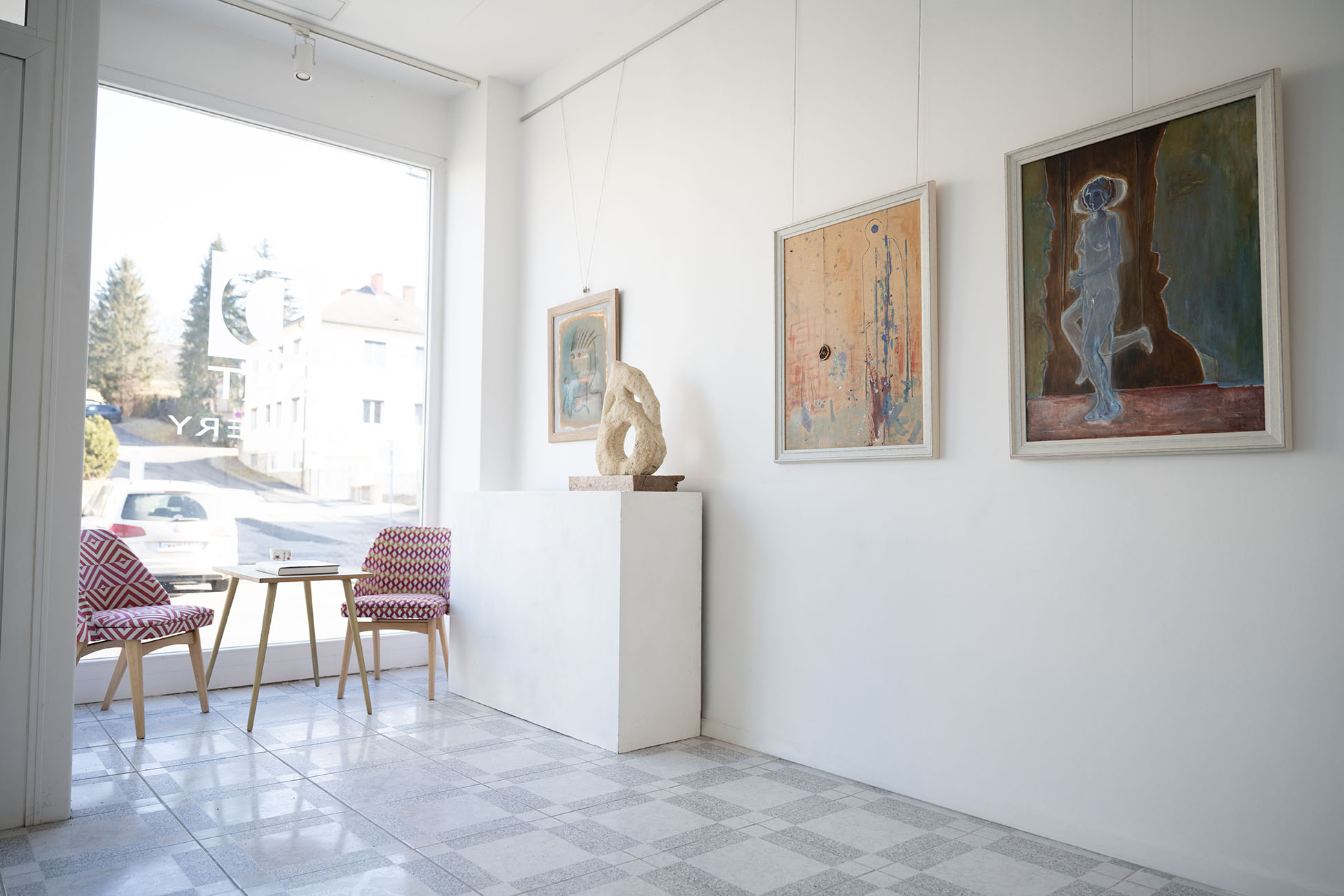 Galeriesitzbereich mit zwei gemusterten Stühlen, kleinem Tisch und Kunstwerken an weißen Wänden während des Tages.