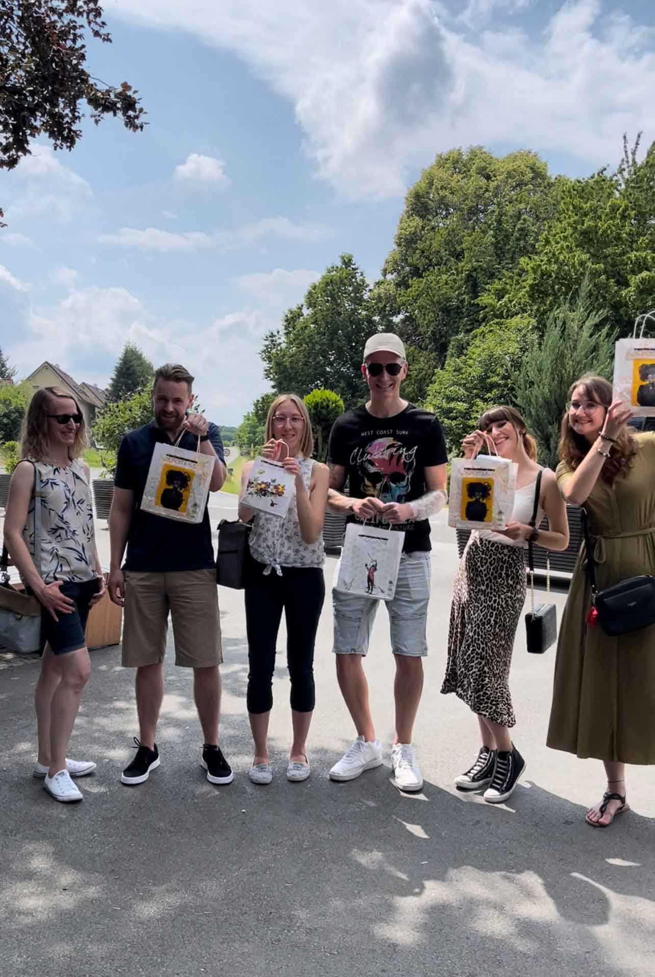 Gruppe von sechs Personen im Freien mit Geschenktüten bei sonnigem Wetter, lächelnd und entspannt.