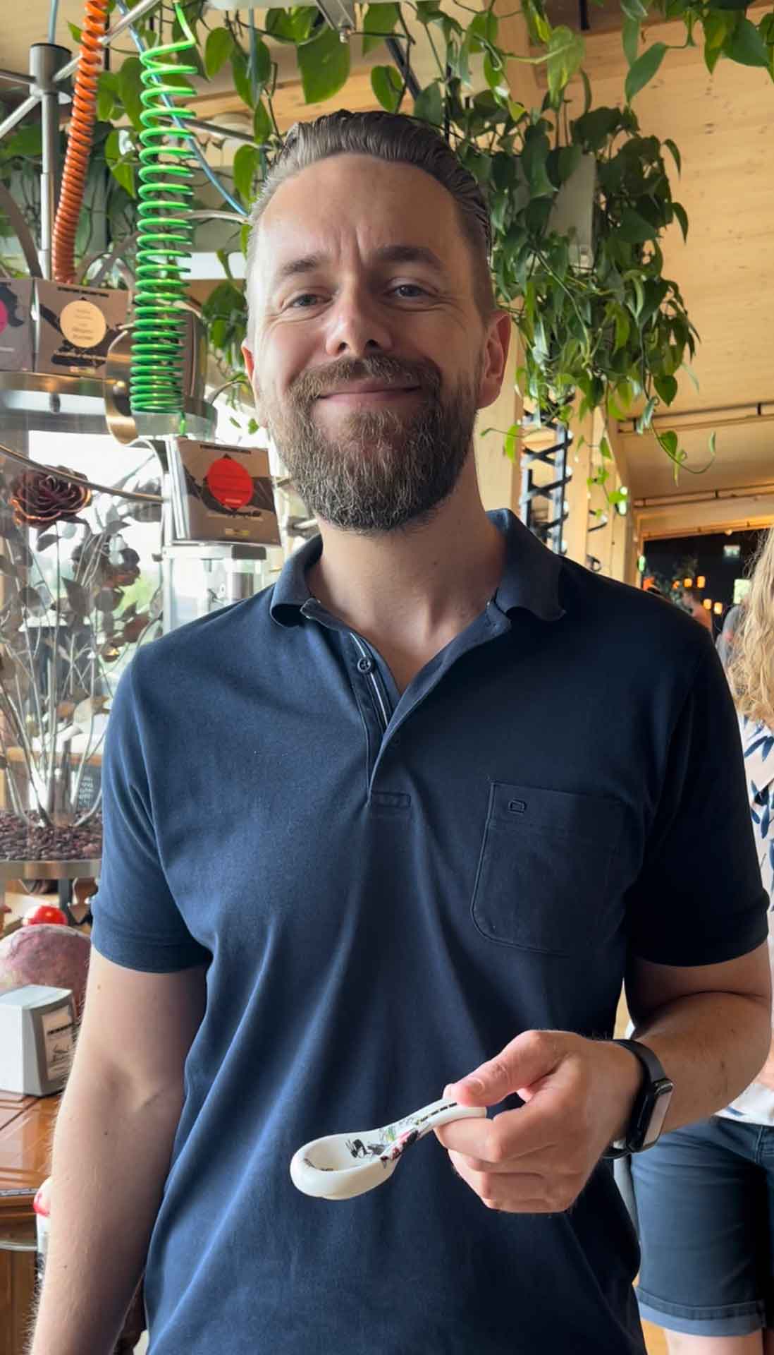 Person hält einen Löffel in einem Raum mit Pflanzen, im Hintergrund moderne Einrichtung.