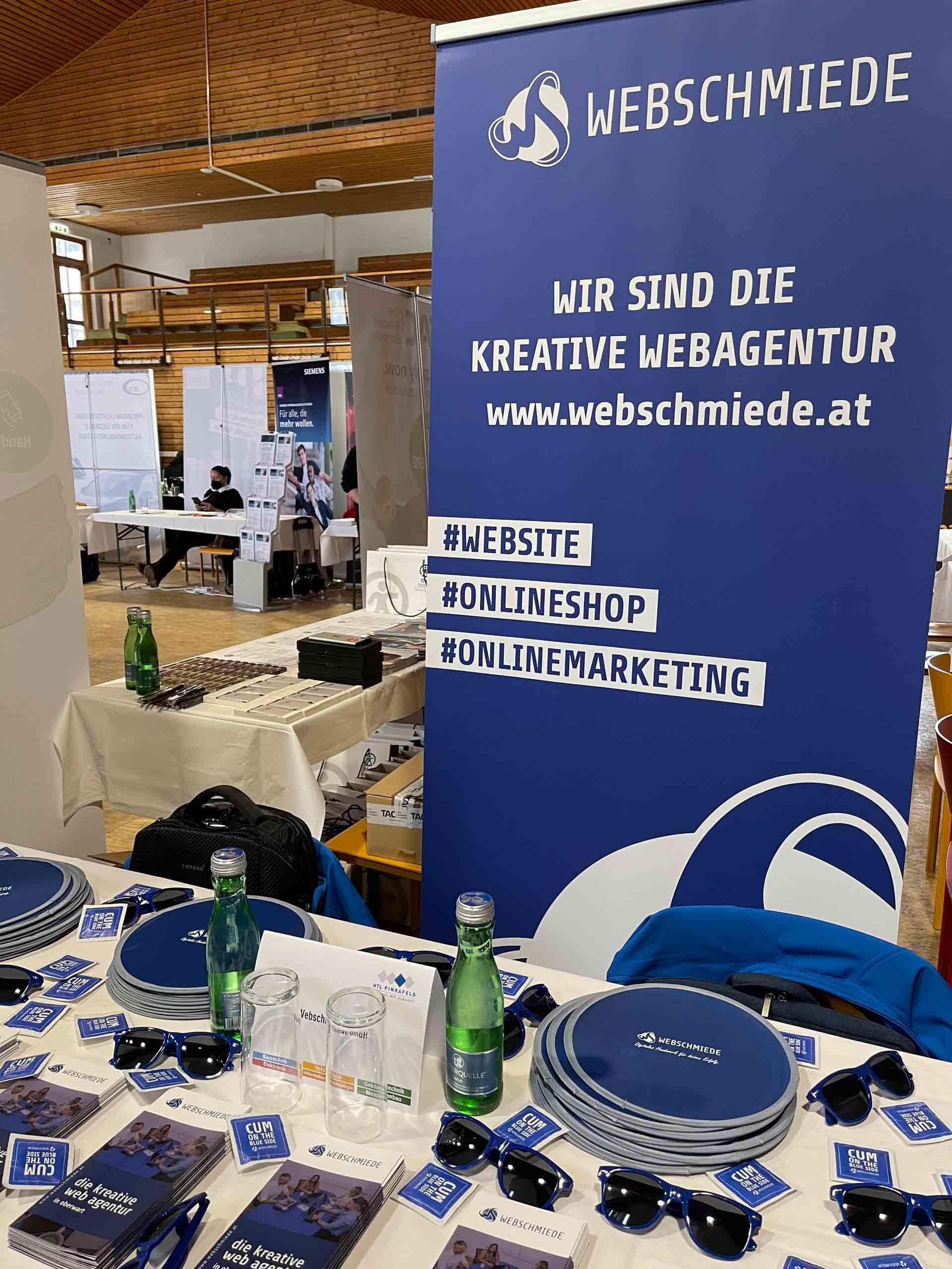 Messestand der Webschmiede mit Bannern, Werbeartikeln und Infomaterial zu Webdesign, SEO und WordPress in einer Halle.