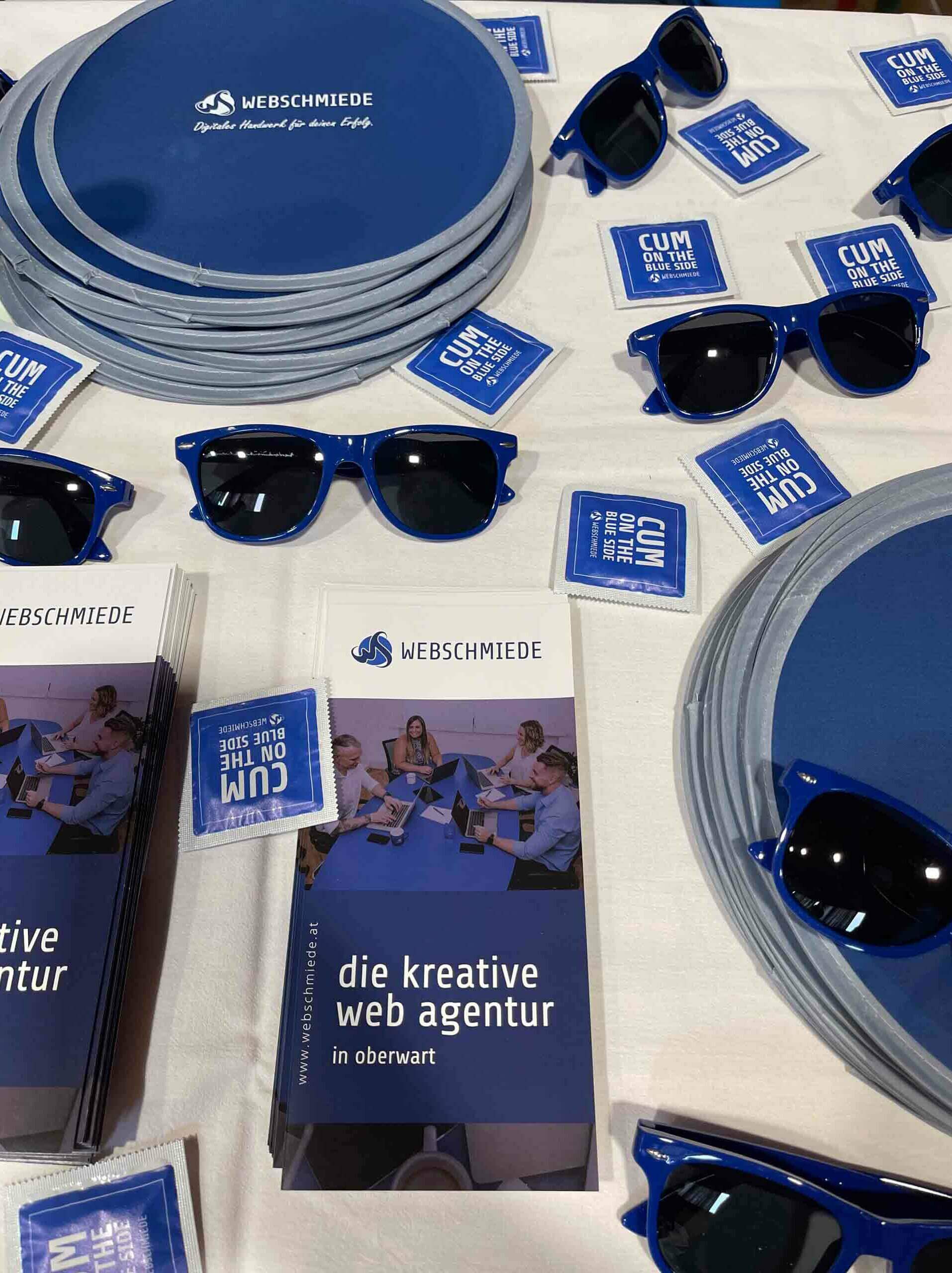Flyer der Webschmiede Agentur mit blauen Sonnenbrillen und Werbematerial auf einem Tisch.