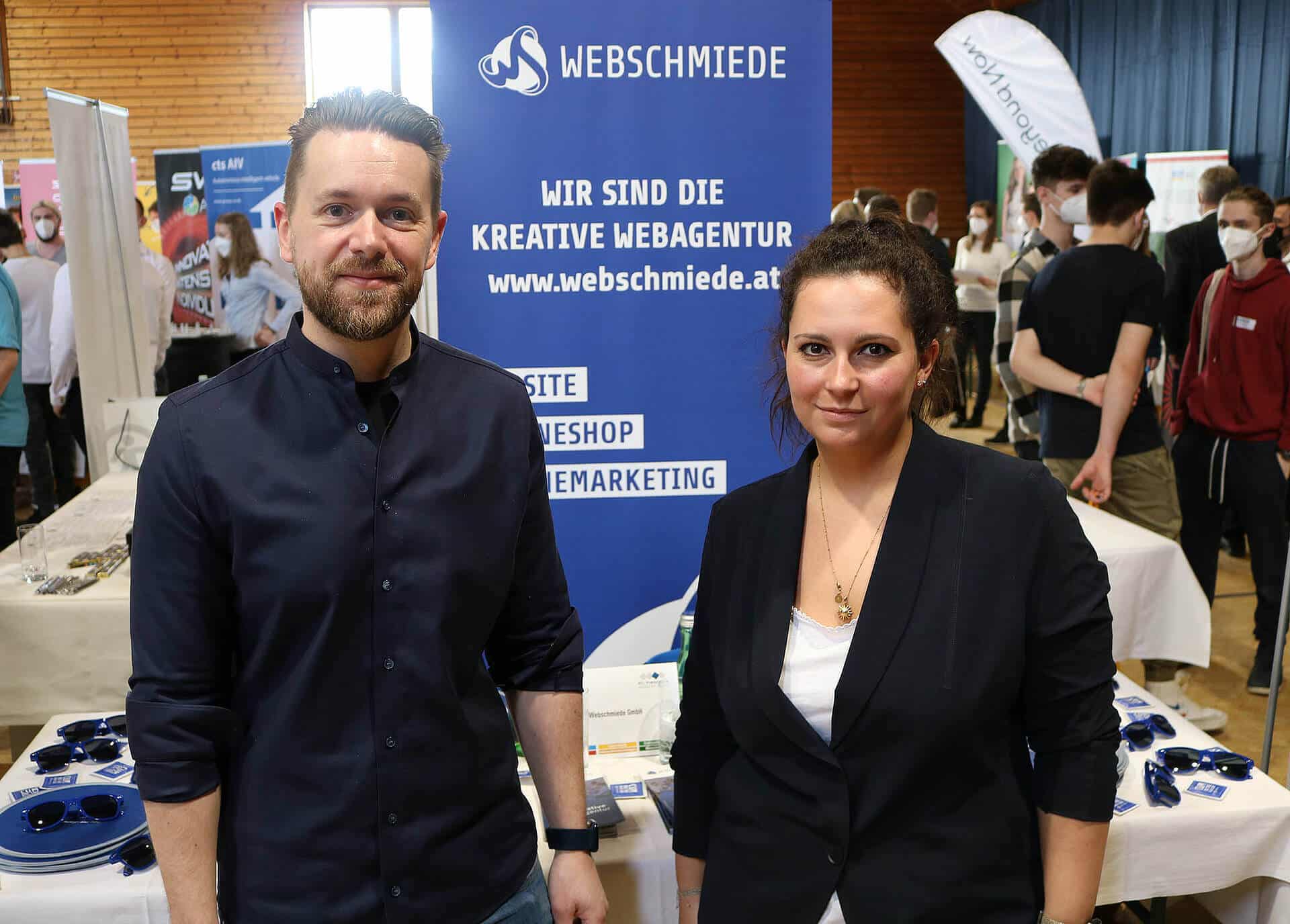 Team der Webschmiede präsentiert WordPress-Dienste auf Messe; Plakat im Hintergrund zeigt Webdesign, SEO und Webshops.