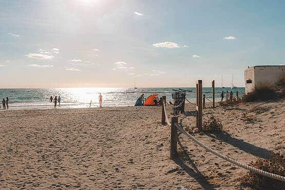 Strand mit Personen, die am Wasser spazieren, Sonnenschirme und ruhige Meereskulisse bei Sonnenuntergang im Hintergrund.