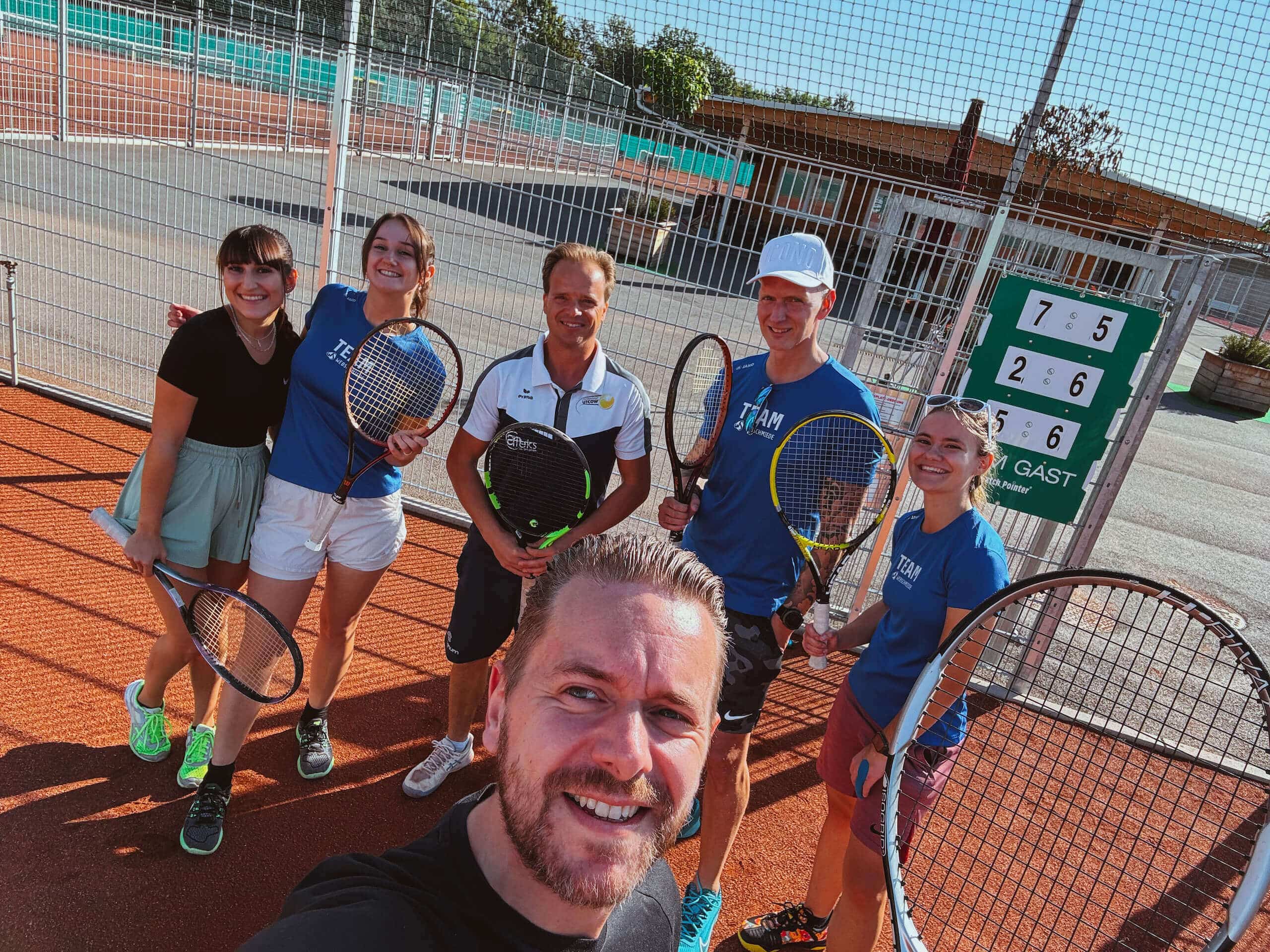 Teambuilding-Event der Webschmiede: Mitarbeiter lächelnd auf dem Tennisplatz, halten Schläger, sonniges Wetter.