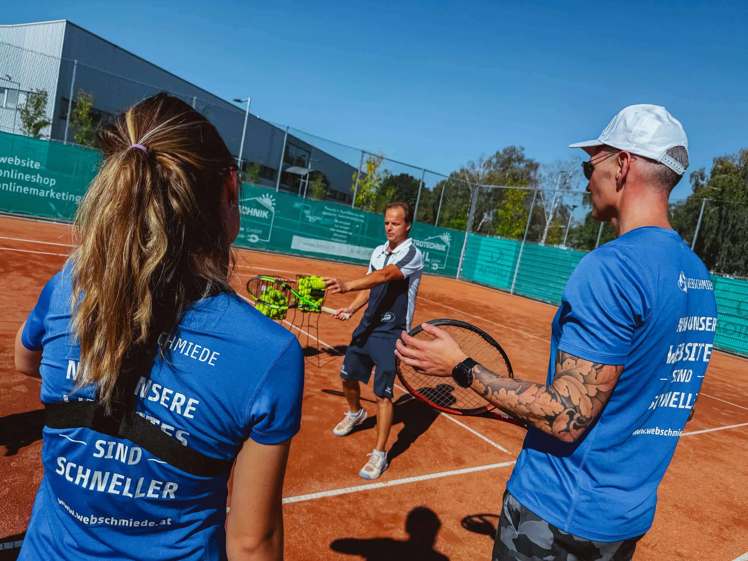 Team in Webschmiede-Shirts spielt Tennis, links Schlägerkorb mit Bällen, sonniger Außenplatz, sportlicher Teamgeist.