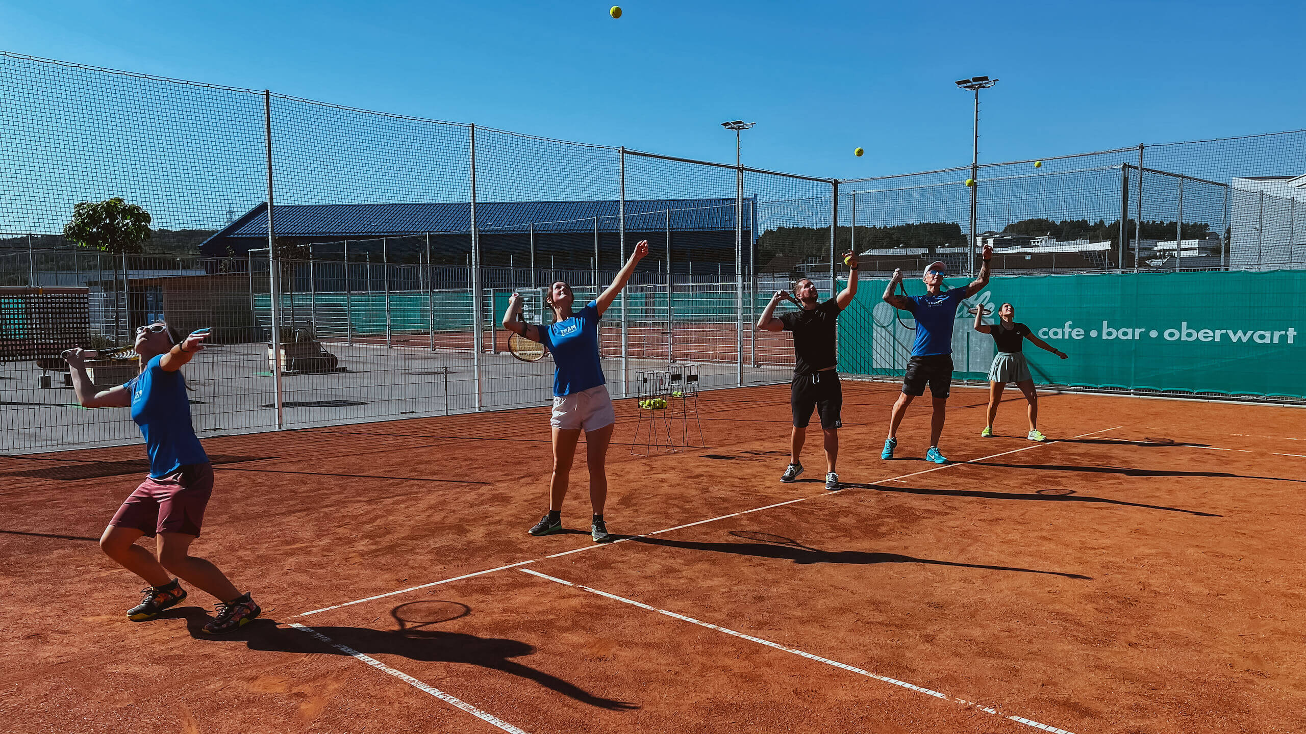 Personengruppe spielt Tennis auf einem Außencourt in Oberwart, mit Bällen und Schlägern, vor blauem Himmel.