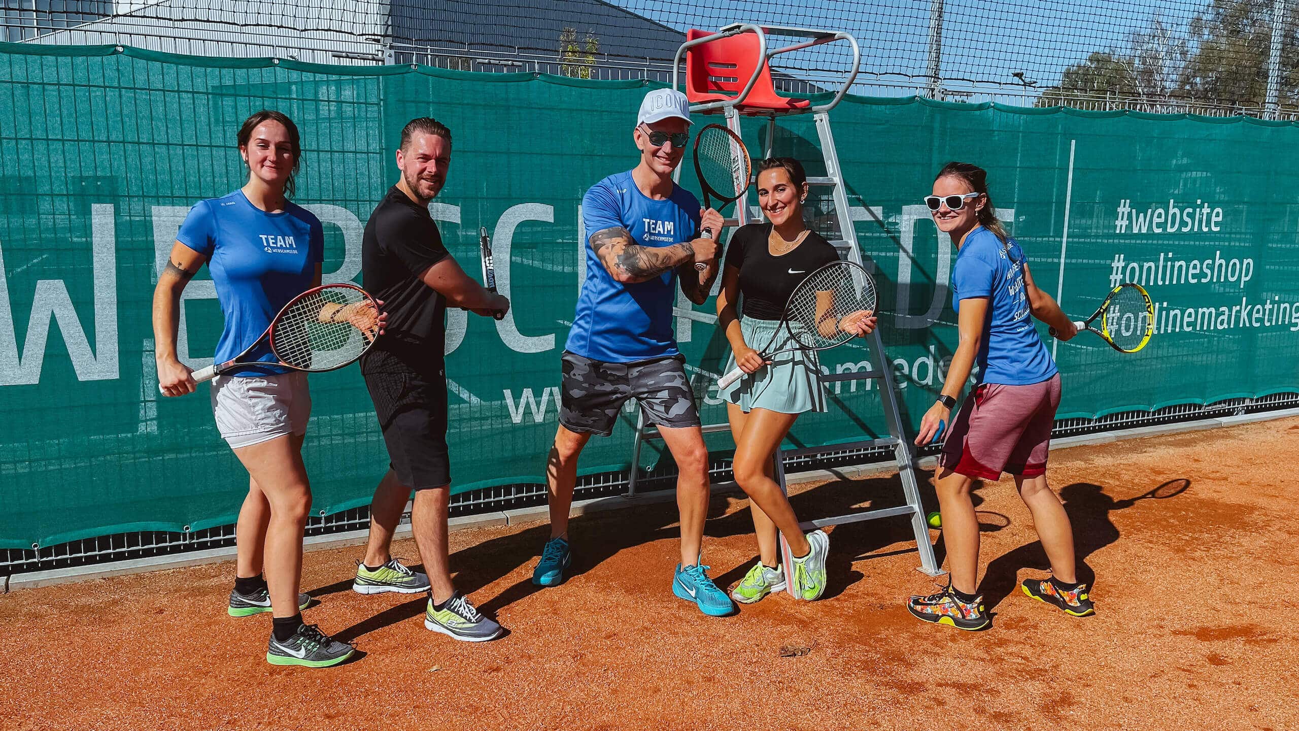 Das Webschmiede-Team spielt Tennis vor einem Banner mit Webdesign- und Online-Marketing-Hashtags.