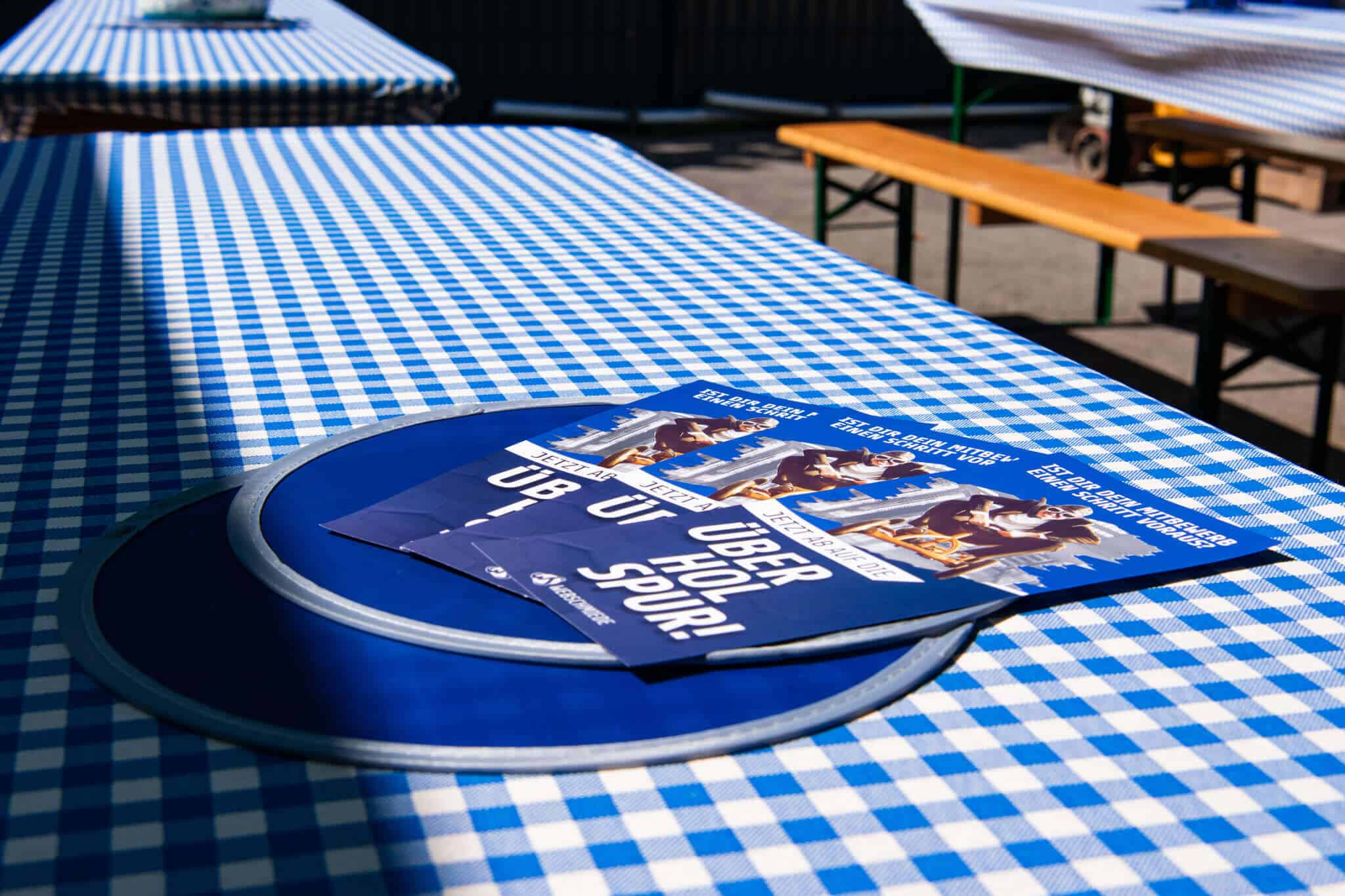 Flyer auf blau-weiß karierter Tischdecke im Biergarten, Fokus auf Event- oder Werbematerial im Freien.