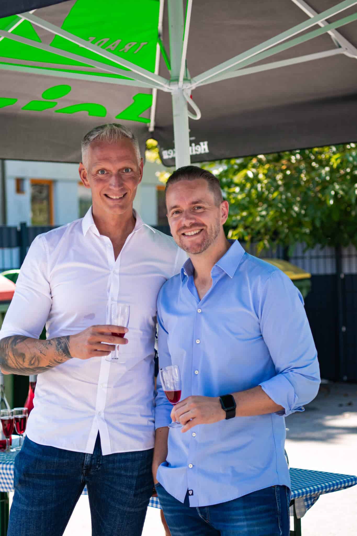 Zwei Personen in Freizeitkleidung bei einem Outdoor-Event, halten Gläser mit Getränken und lächeln in die Kamera.