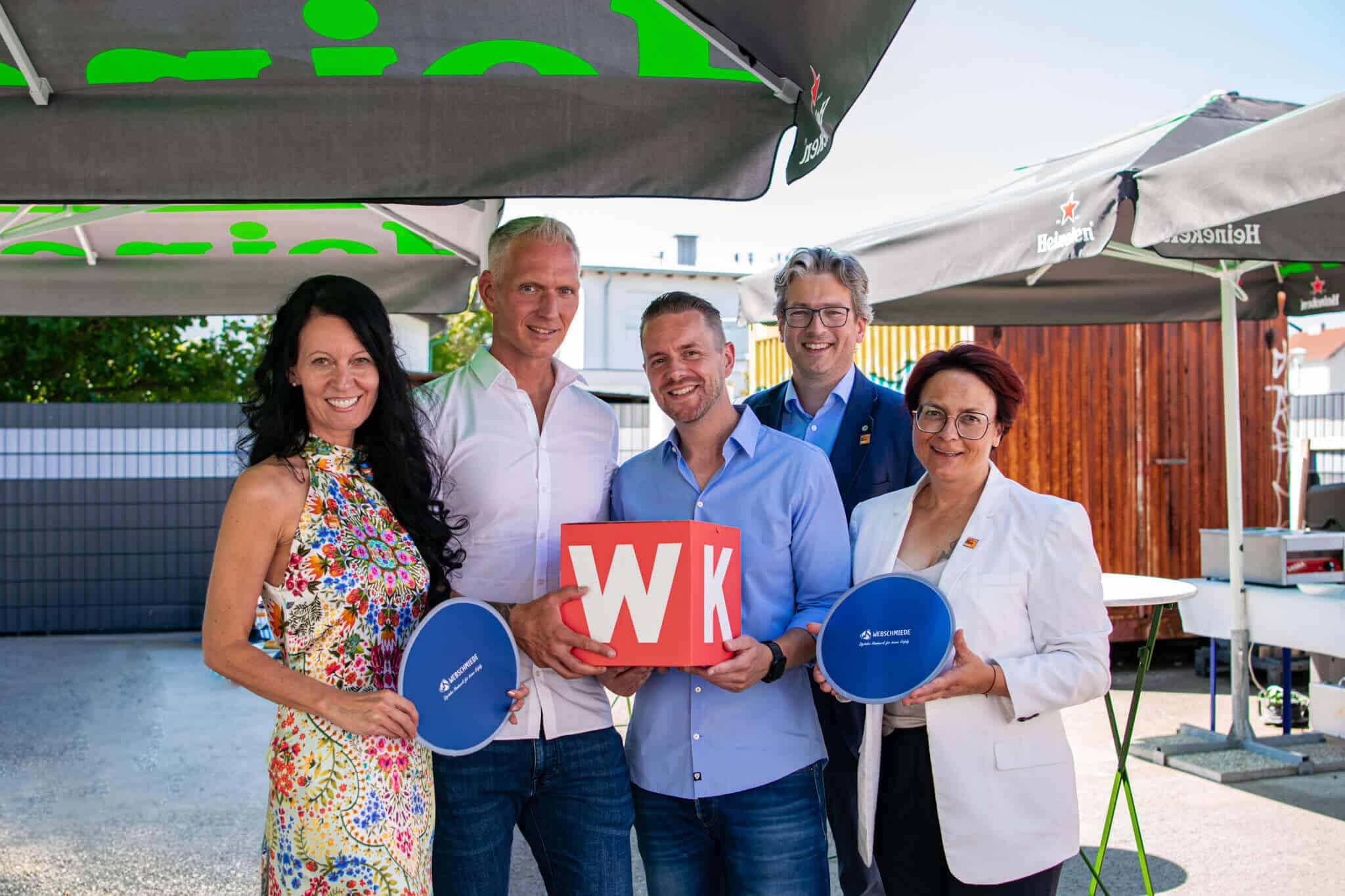 Gruppe von fünf Personen posiert draußen mit einem Würfel und Schildern von Webschmiede unter Sonnenschirmen.