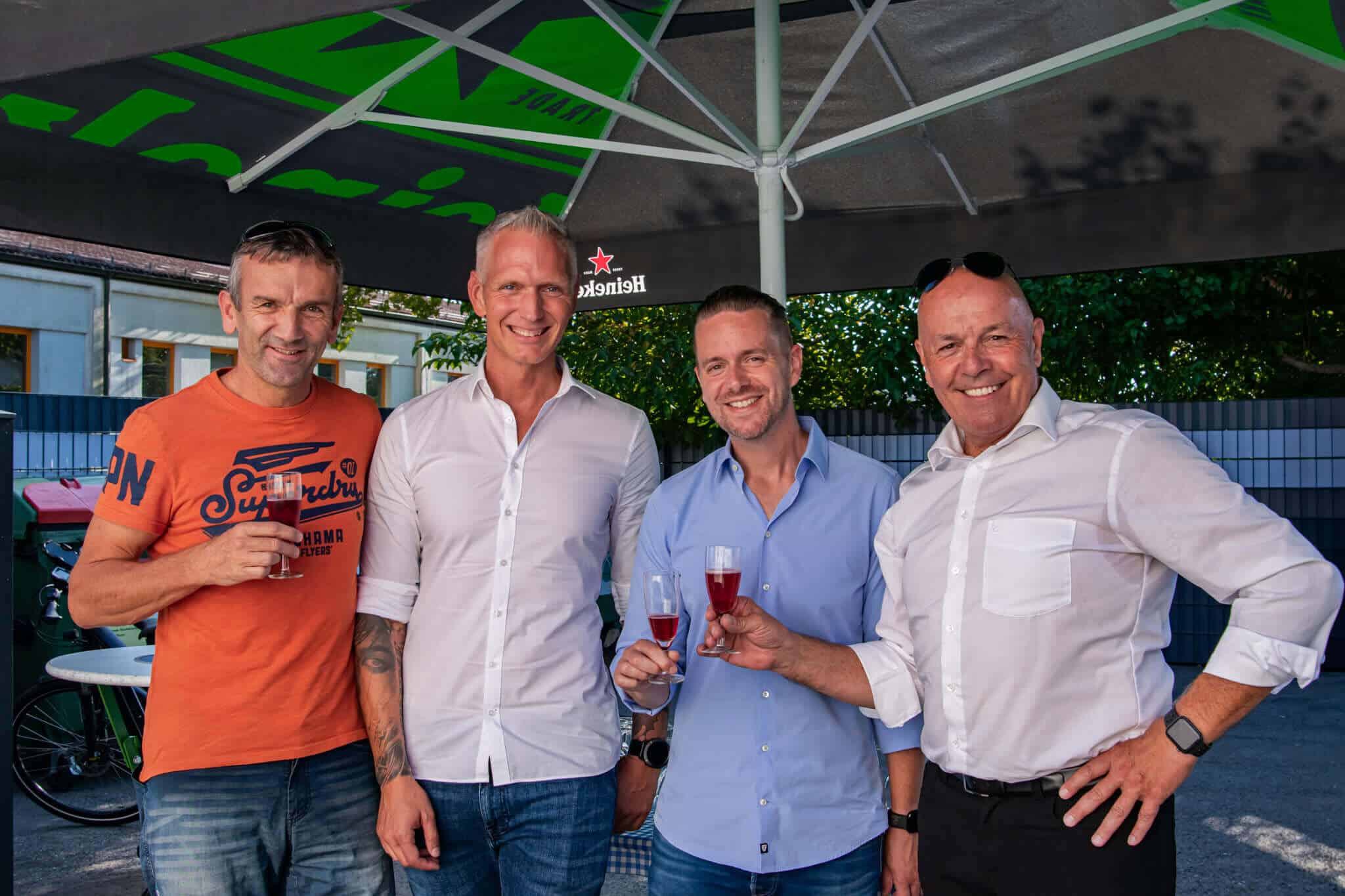 Vier Männer lächeln unter einem Sonnenschirm, stoßen mit Gläsern im Freien an, lockere Atmosphäre.
