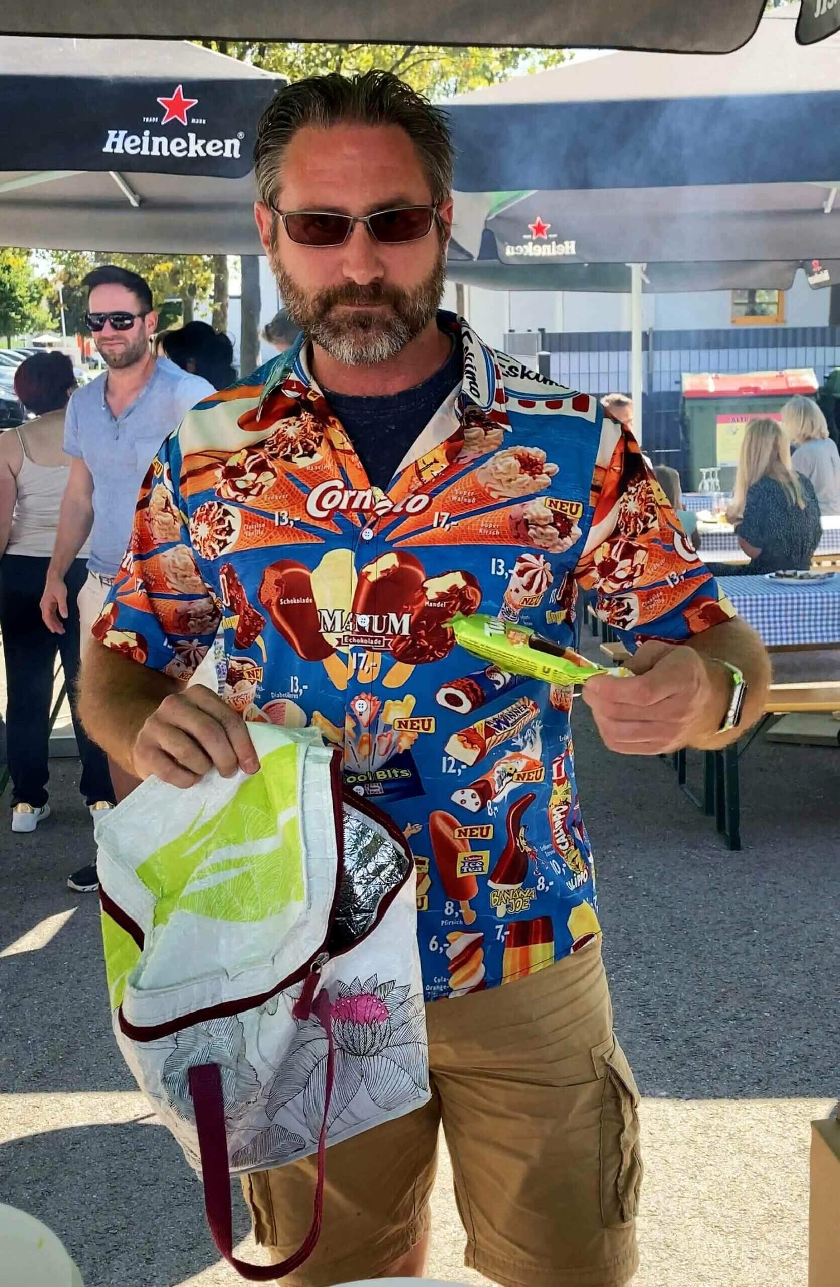 Person in bunter Eiskrem-Hemd bei sommerlichem Event, umgeben von Menschen unter Schirmen im Freien.