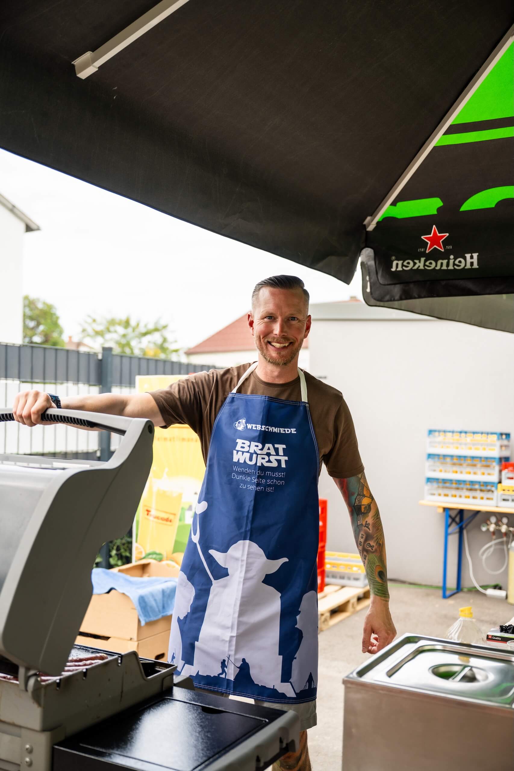 Person in Webschmiede-Schürze grillt Bratwürste bei einem Outdoor-Event.