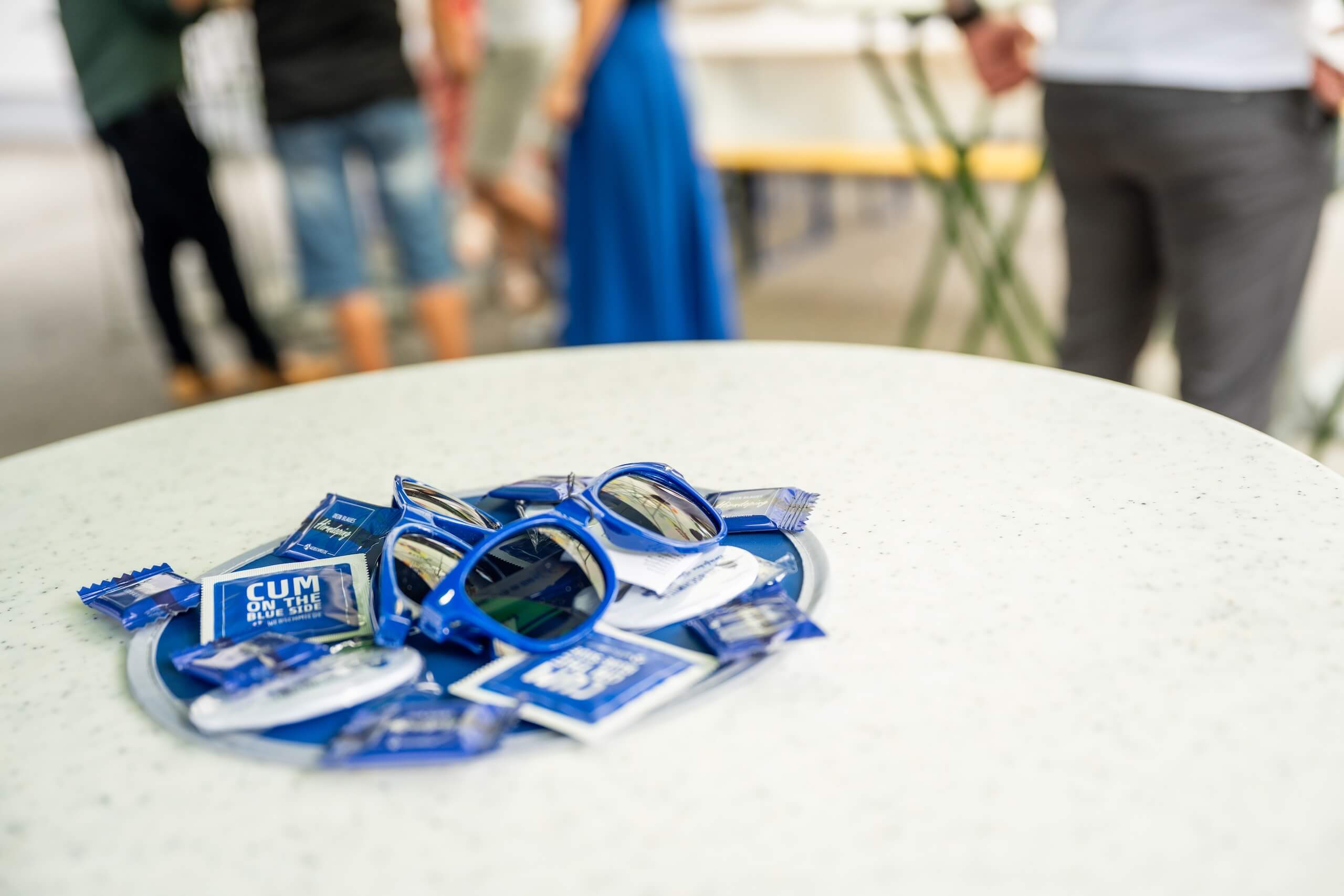 Blaue Sonnenbrille und Werbematerial der Webschmiede auf einem Tisch, unscharfe Personen im Hintergrund, Networking-Event.