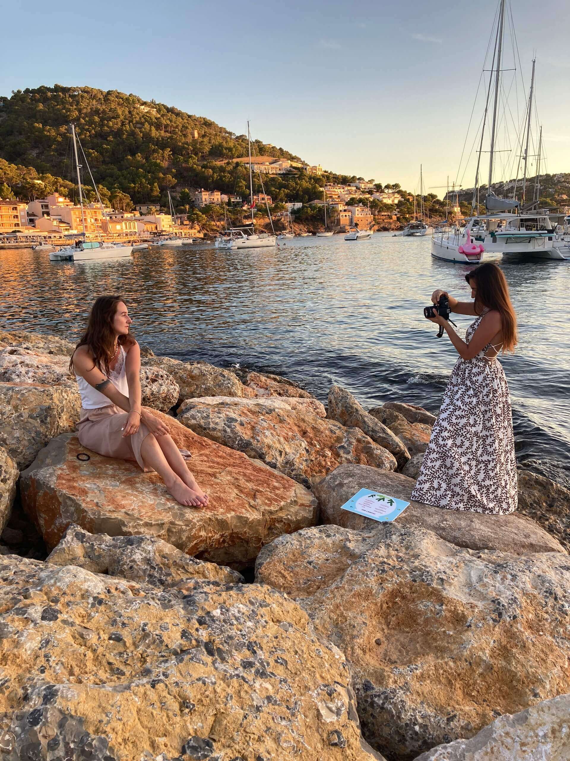 Eine Person fotografiert eine andere, die auf Felsen am Hafen mit Segelbooten im Hintergrund sitzt.