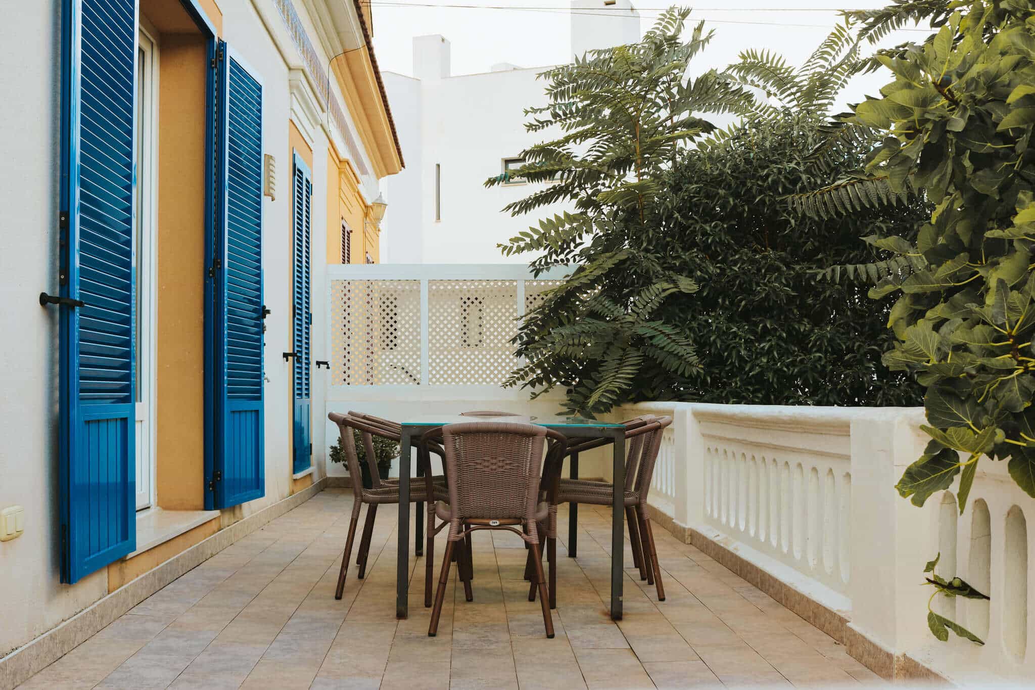 Terrasse mit Tisch und Stühlen, umgeben von mediterranen Pflanzen, neben Häusern mit blauen Fensterläden.