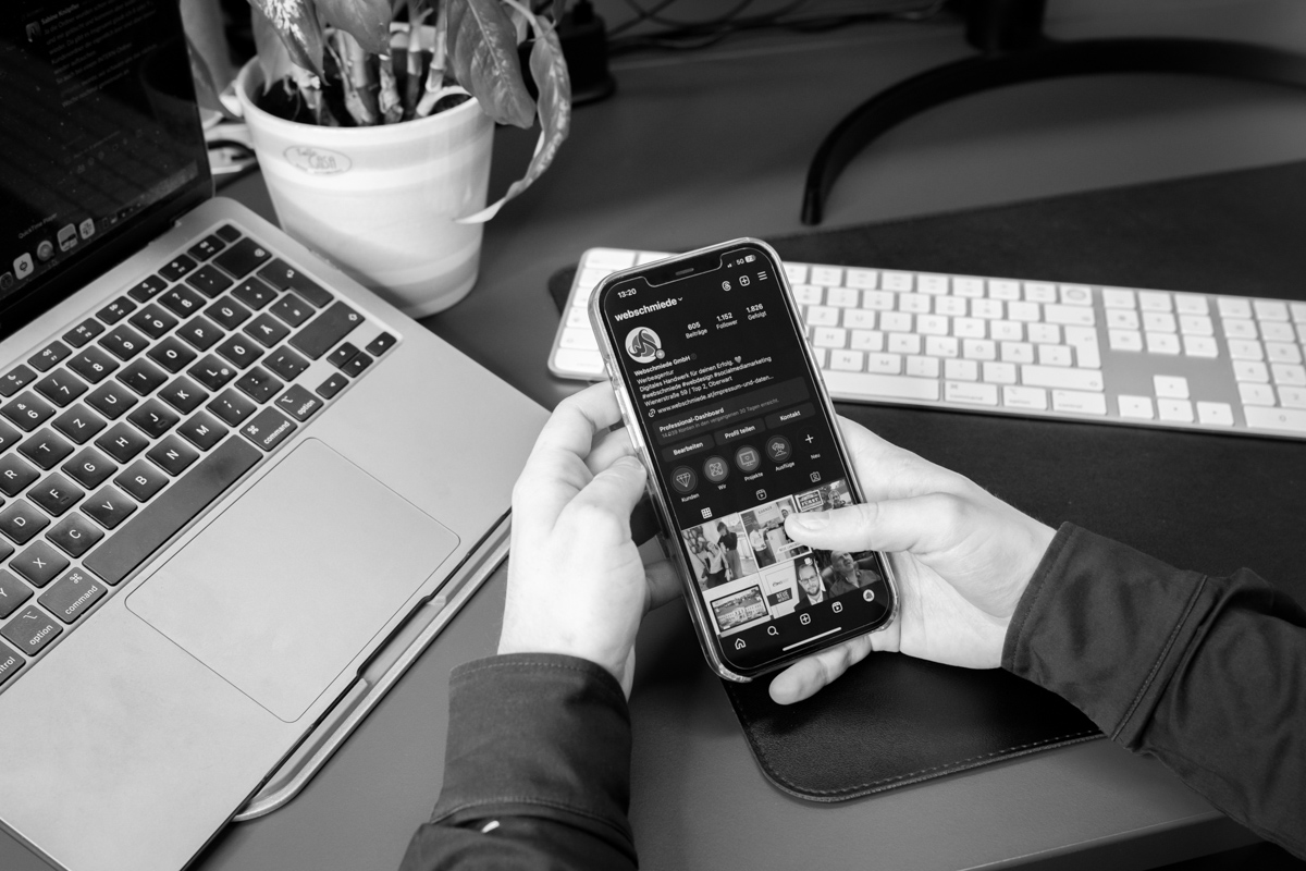 Eine Person hält ein Smartphone mit Instagram-Profil der Webschmiede neben einem Laptop in einem Büro.