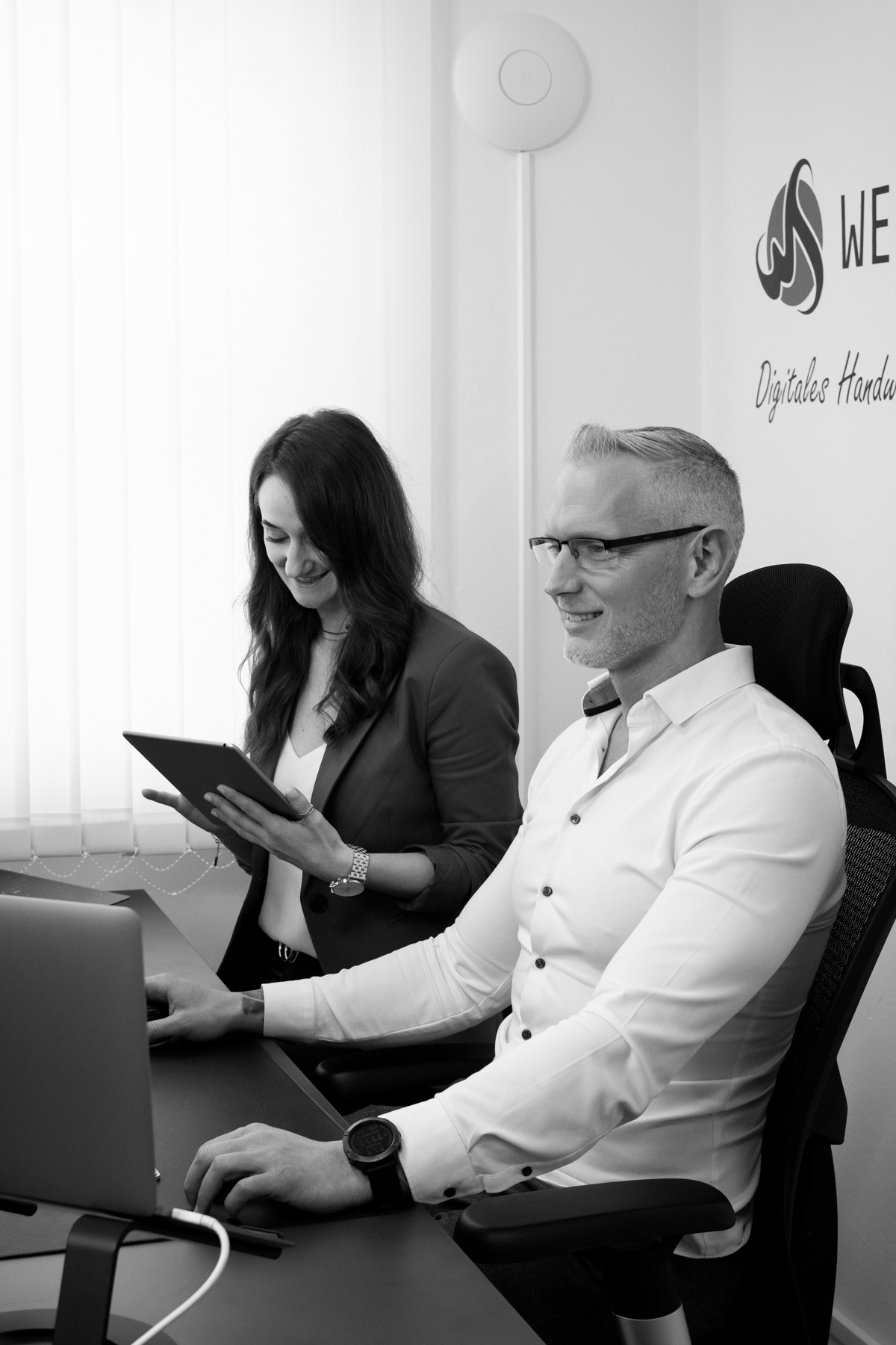 Team von Webschmiede arbeitet zusammen im Büro, eine Person am Laptop, eine mit Tablet, die Wand zeigt das Agentur-Logo.
