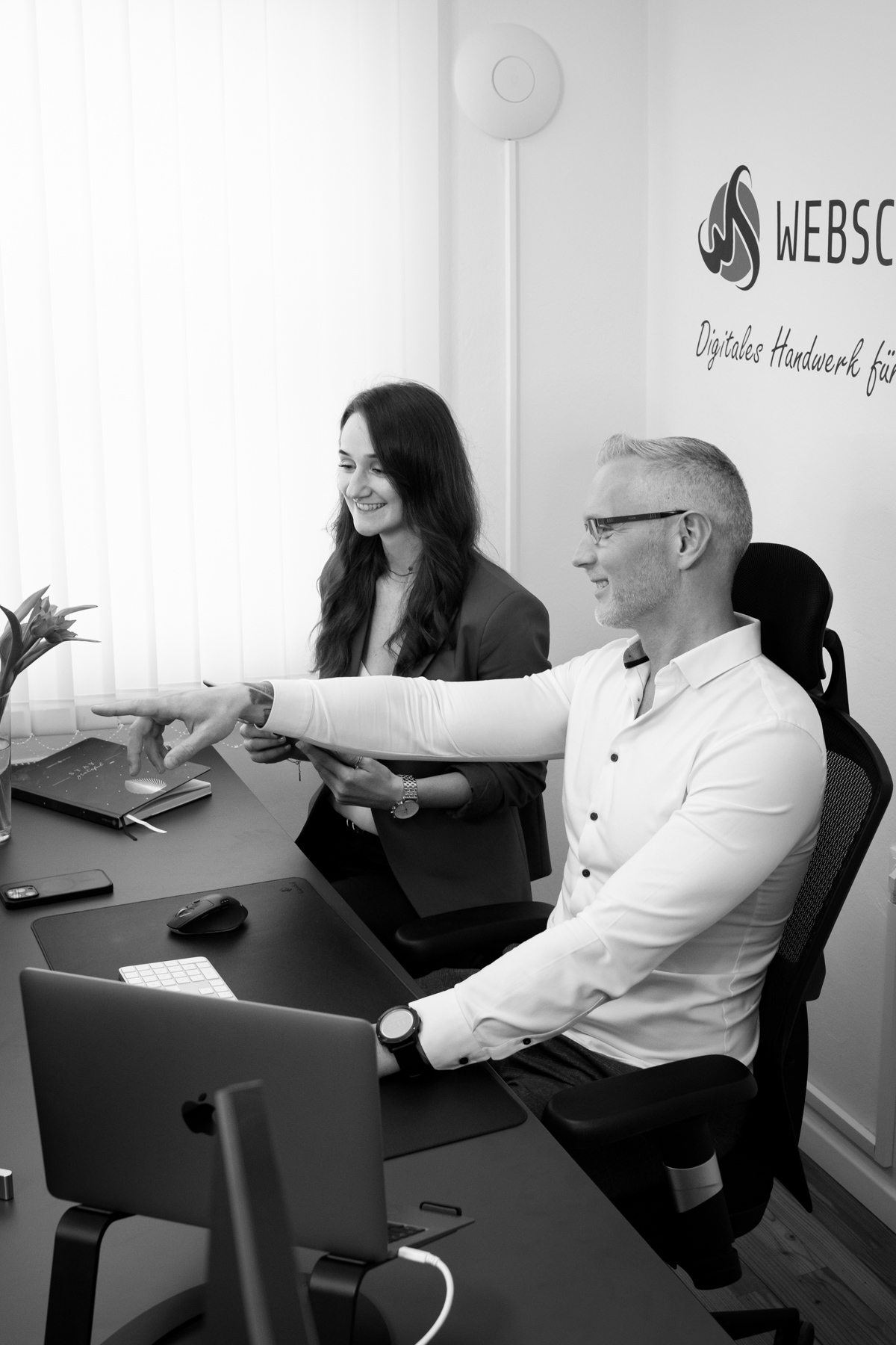 Zwei Personen arbeiten lächelnd an einem Schreibtisch in der Webschmiede WordPress-Agentur, Oberwart.