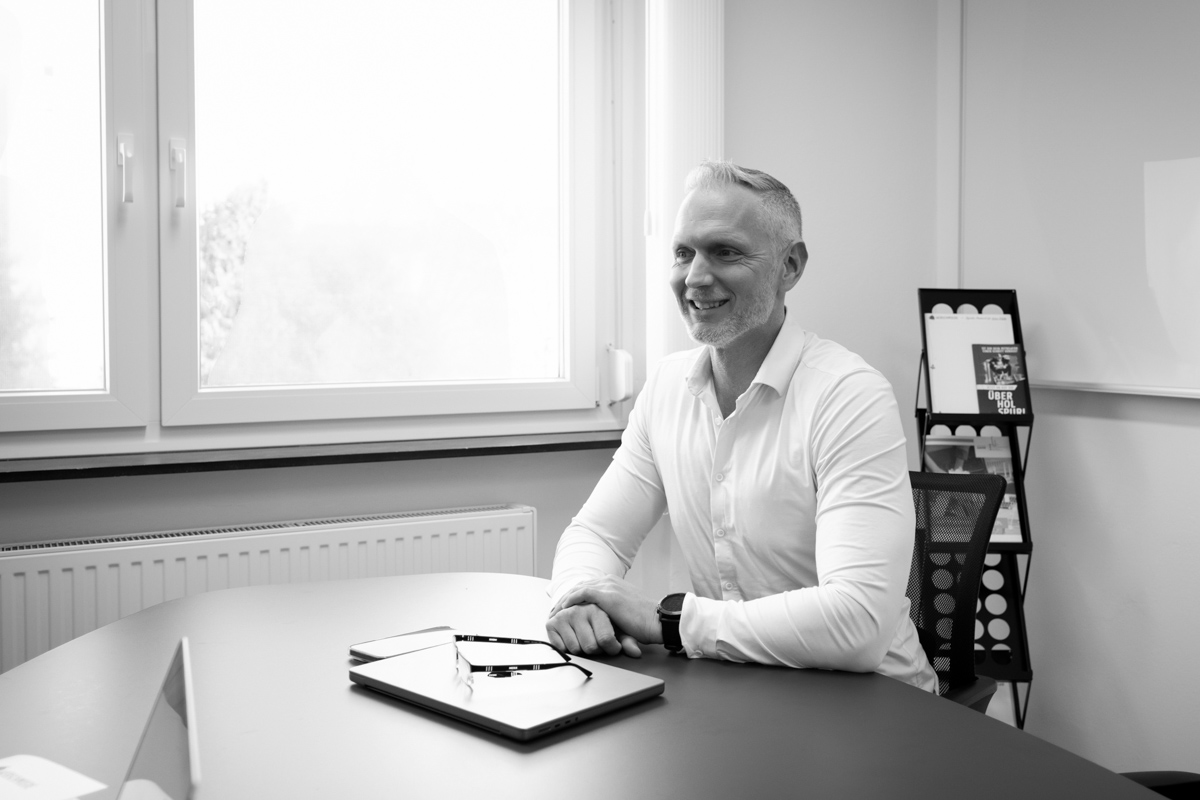 Eine Person in einem Büro, lächelt am Tisch, mit Laptop und Brille, im Hintergrund Zeitschriftenständer.