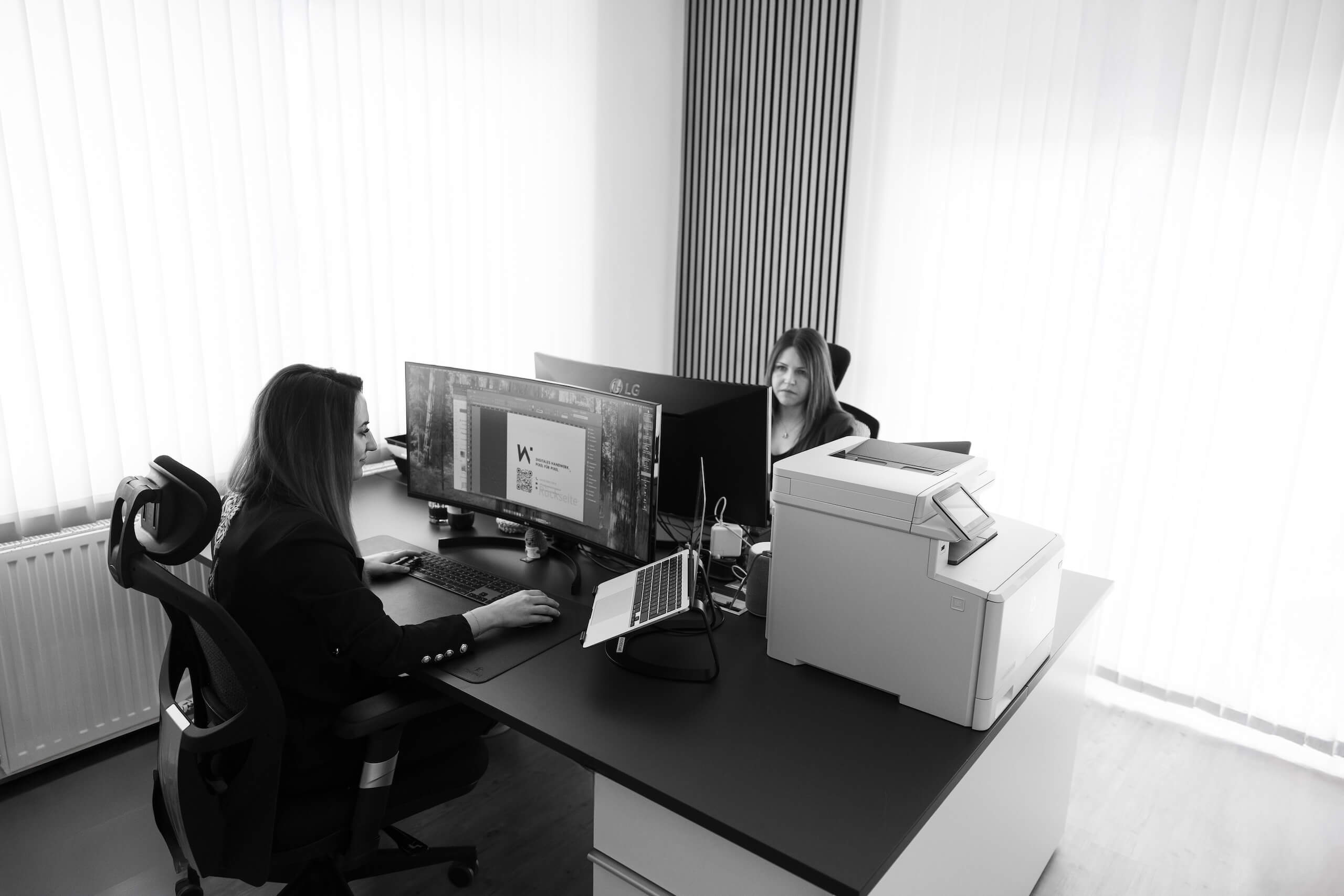 Zwei Mitarbeiterinnen der Webschmiede Agentur arbeiten an modernen Computern in einem Büro, ein Drucker ist im Vordergrund.
