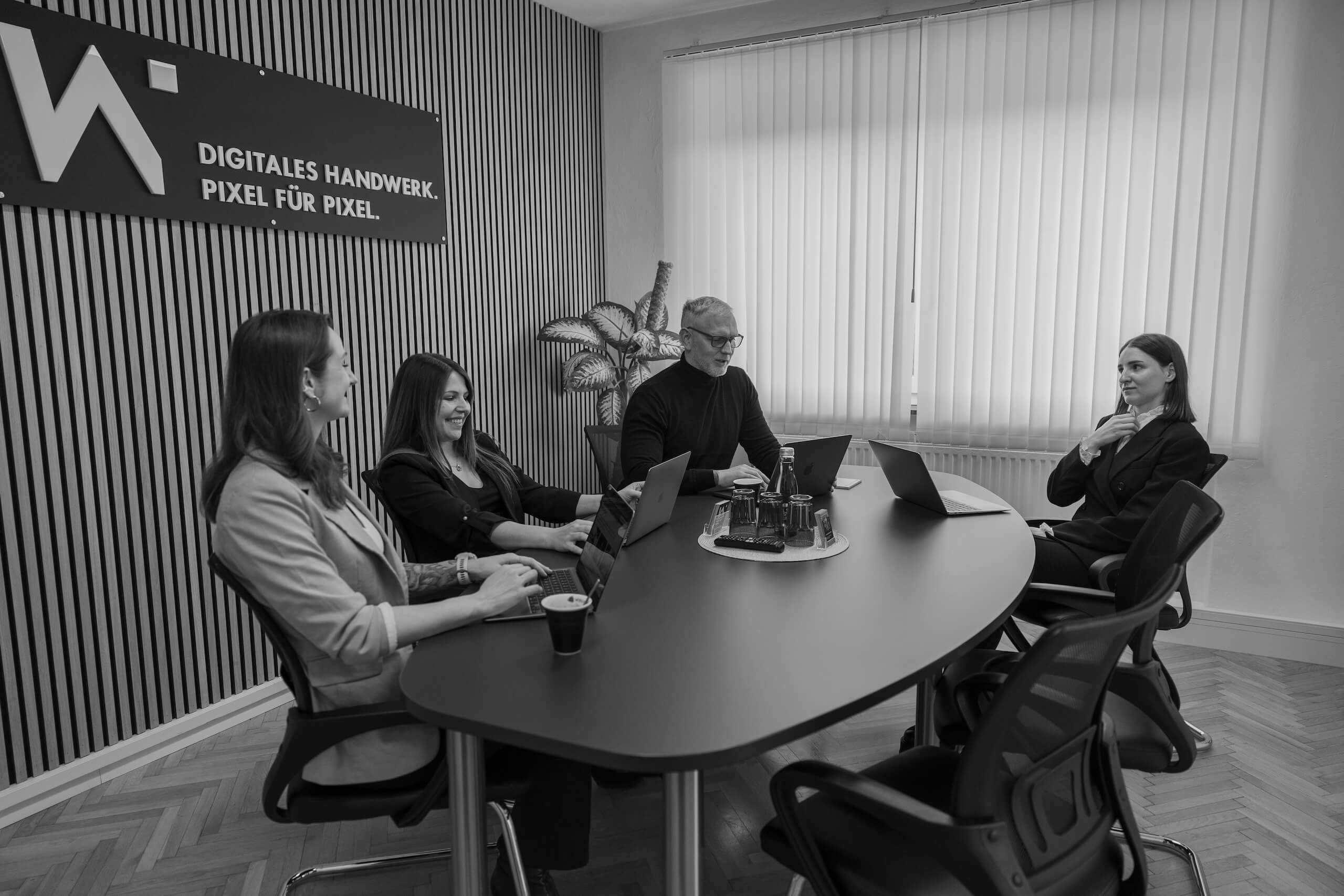 Team der Webschmiede bespricht Webdesign-Projekte an einem Tisch, arbeiten an Laptops, im Büro mit Firmenschild.