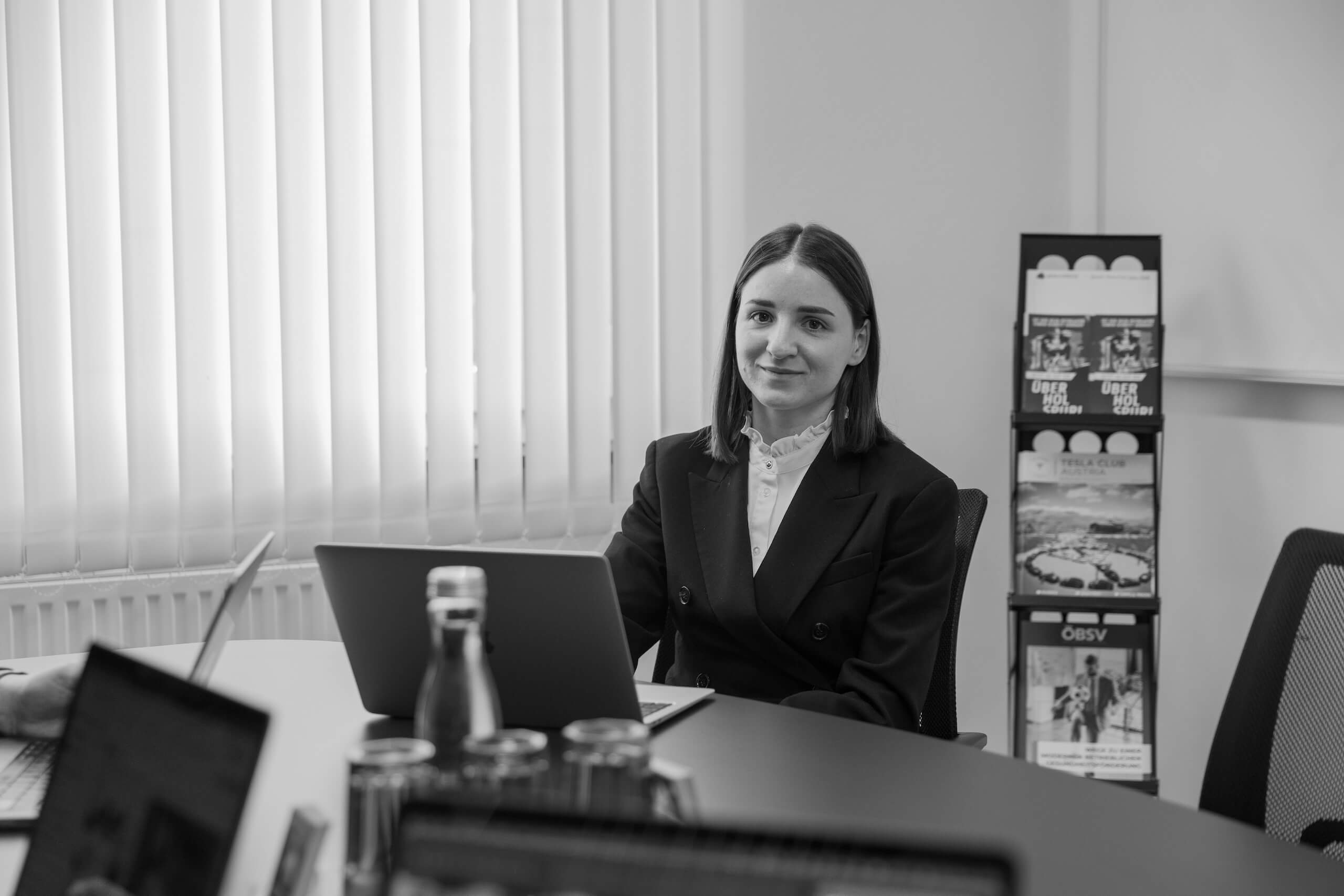 Mitarbeiterin der Webschmiede WordPress Agentur sitzt lächelnd am Schreibtisch mit Laptop und Broschüren im Hintergrund.