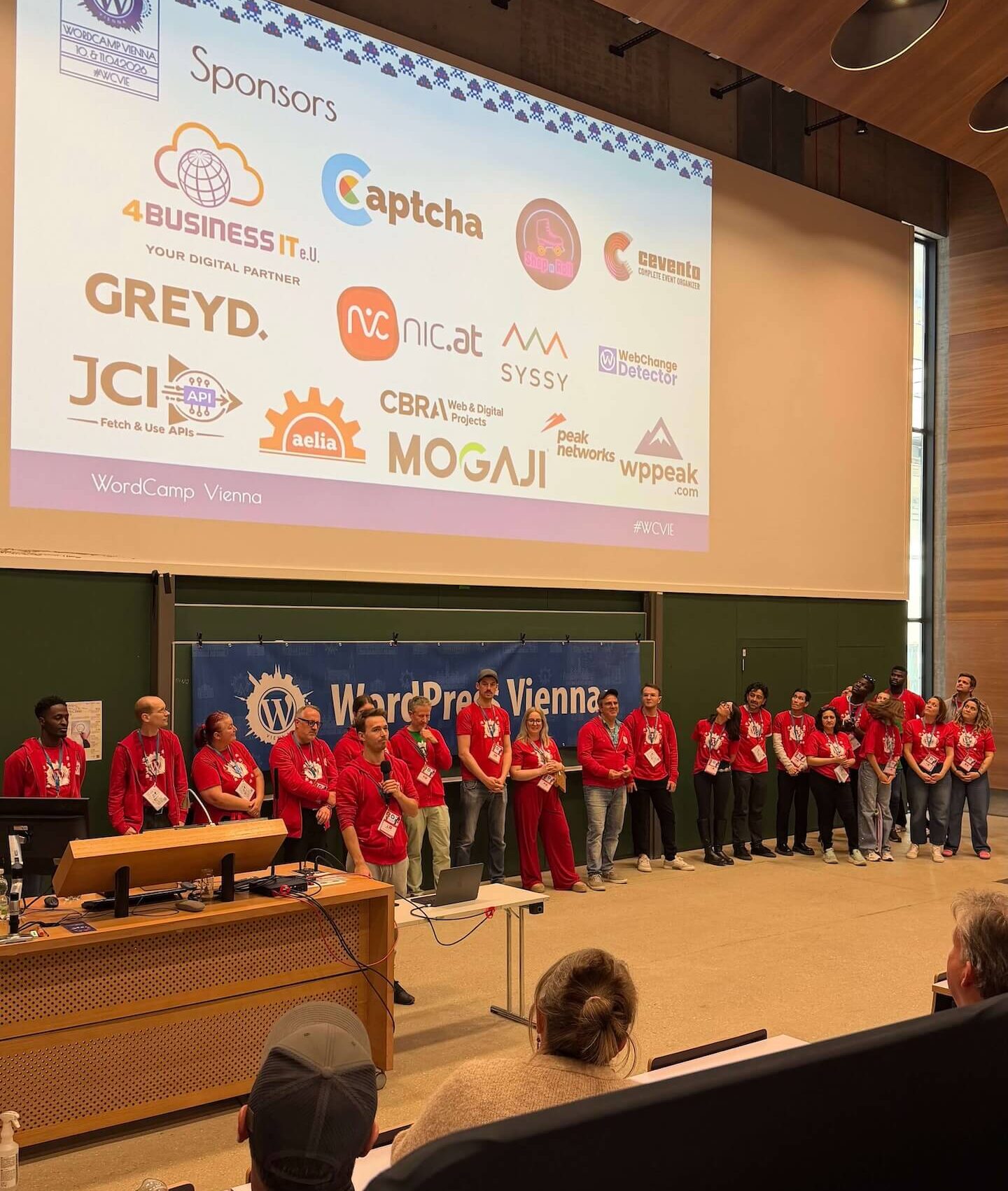 WordCamp Vienna: Menschen in roten T-Shirts vor Sponsorenwand auf Bühne, Web- und WordPress-Event im Konferenzsaal.