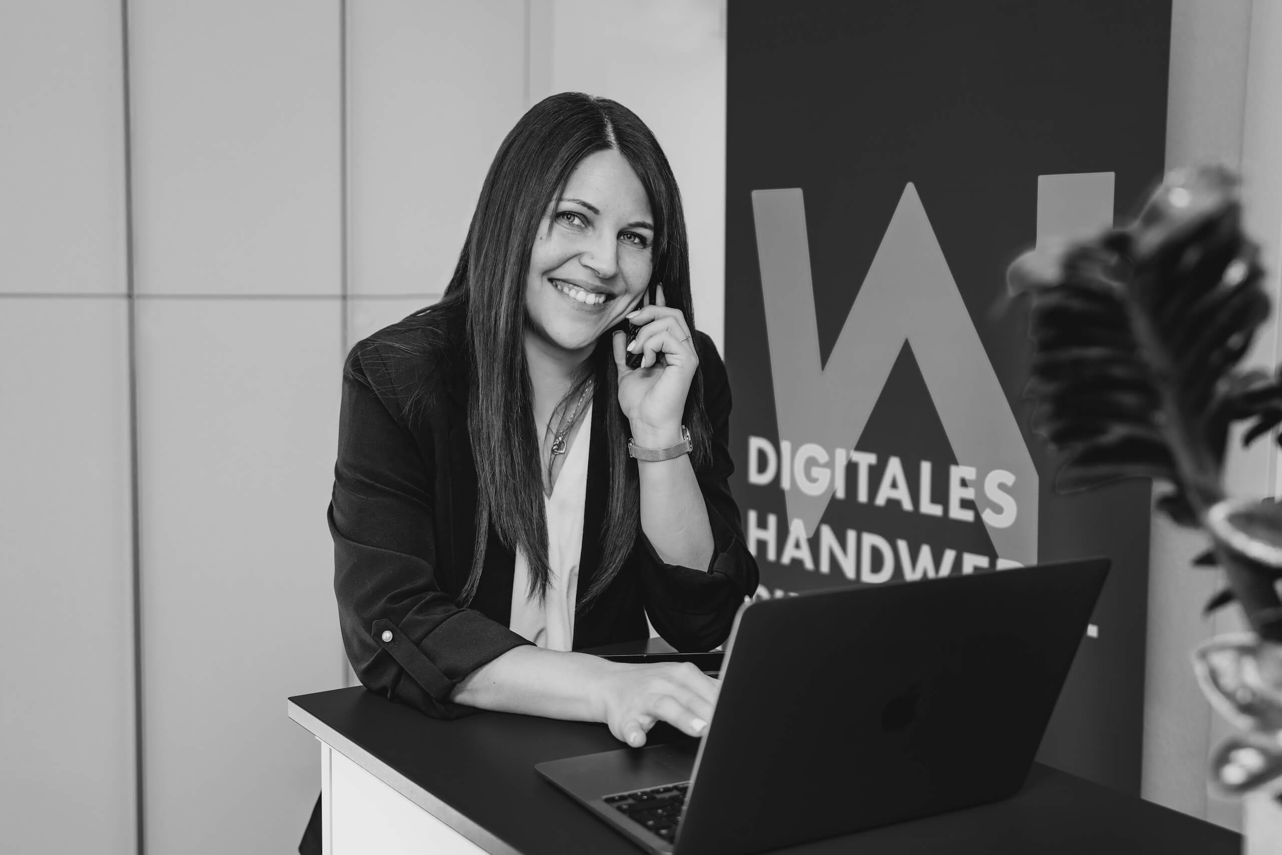 Mitarbeiterin der Webschmiede am Laptop und Handy, lächelnd vor einem Banner mit „Digitales Handwerk“.