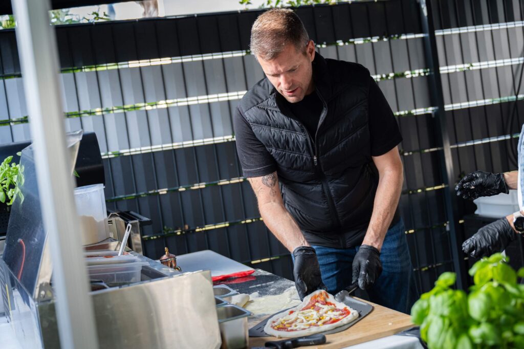 Eine Person bereitet im Freien Pizza zu, umgeben von Zutaten und einem Pizzaofen.