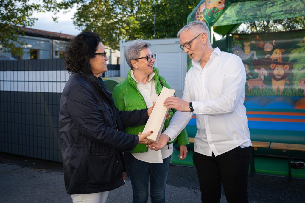 Drei Personen im Gespräch im Freien, eine hält ein Dokument, bunte Hüpfburg im Hintergrund.